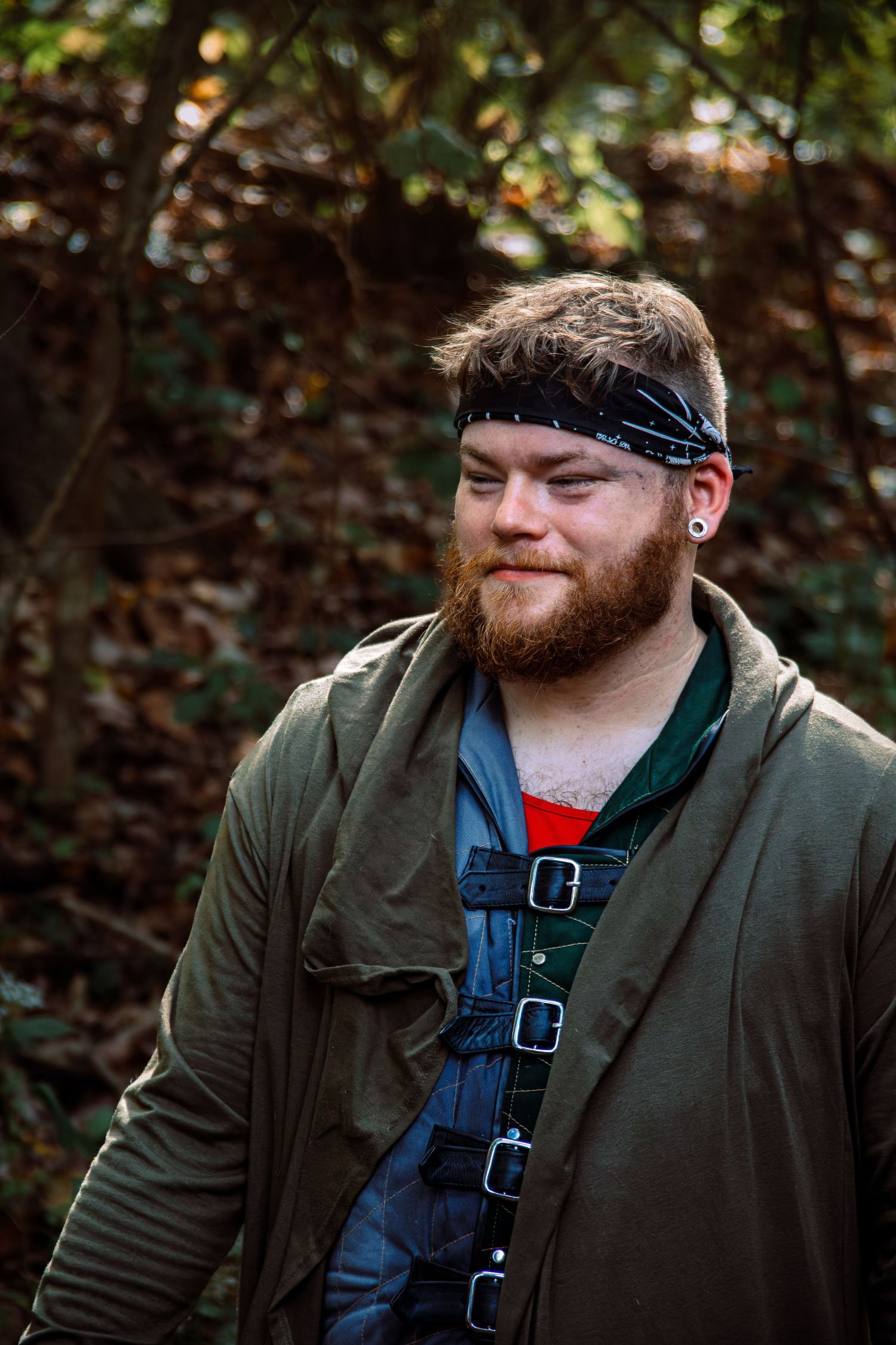 Man with red beard wearing olive jacket and denim overalls poses in outdoor setting.