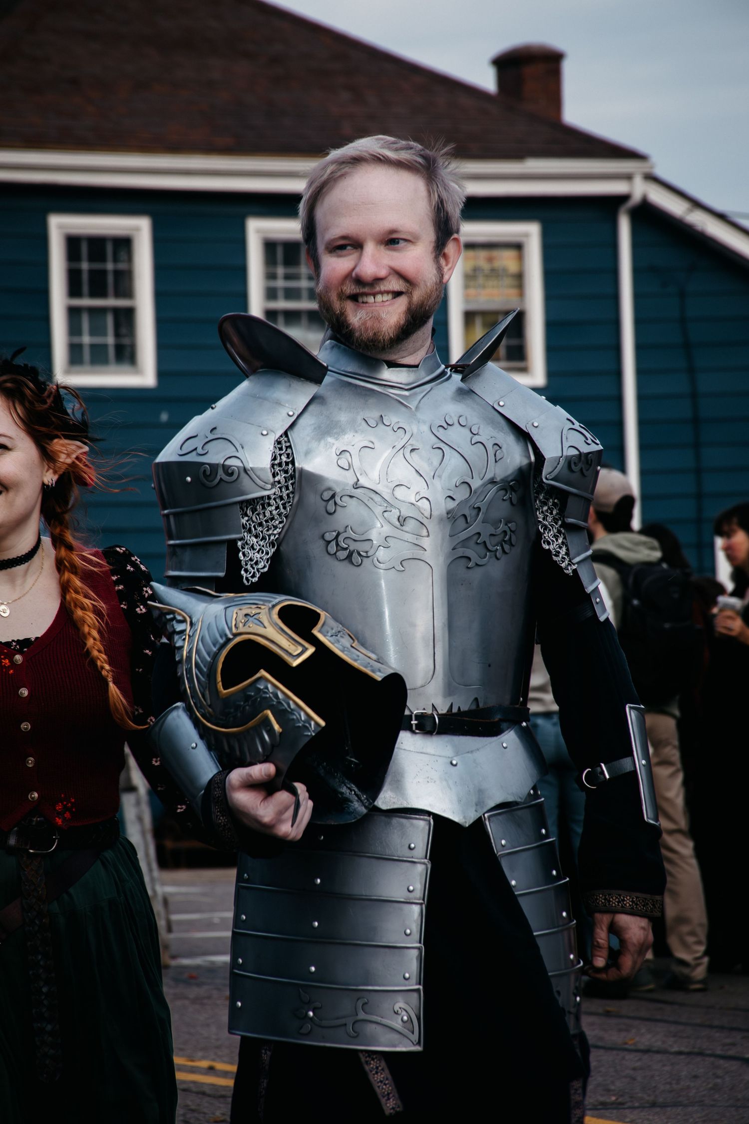 Medieval knight in ornate silver armor with decorative engravings stands outside a blue house during a cosplay event.