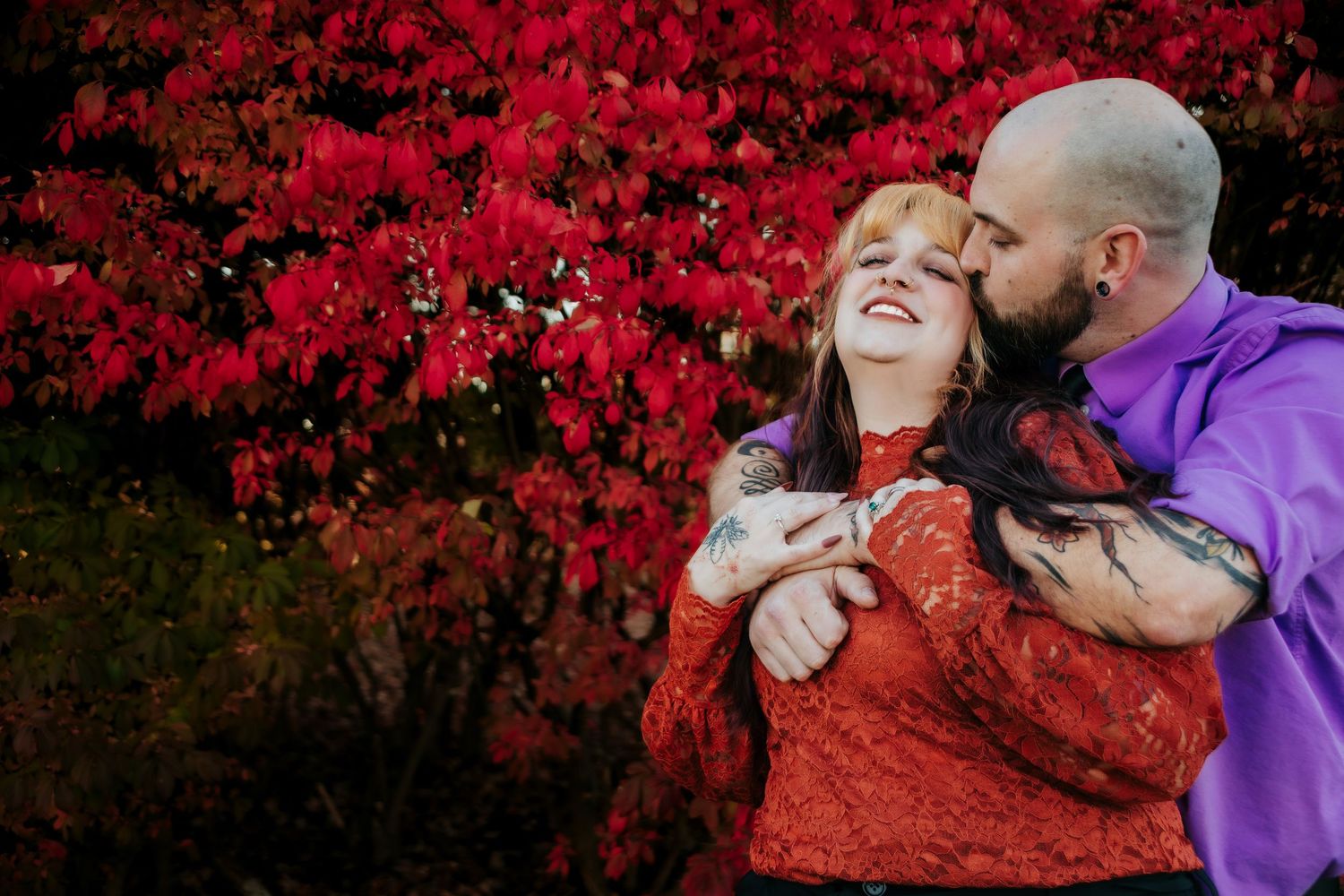 A couple embraces against vibrant red fall foliage creating a romantic autumn scene.