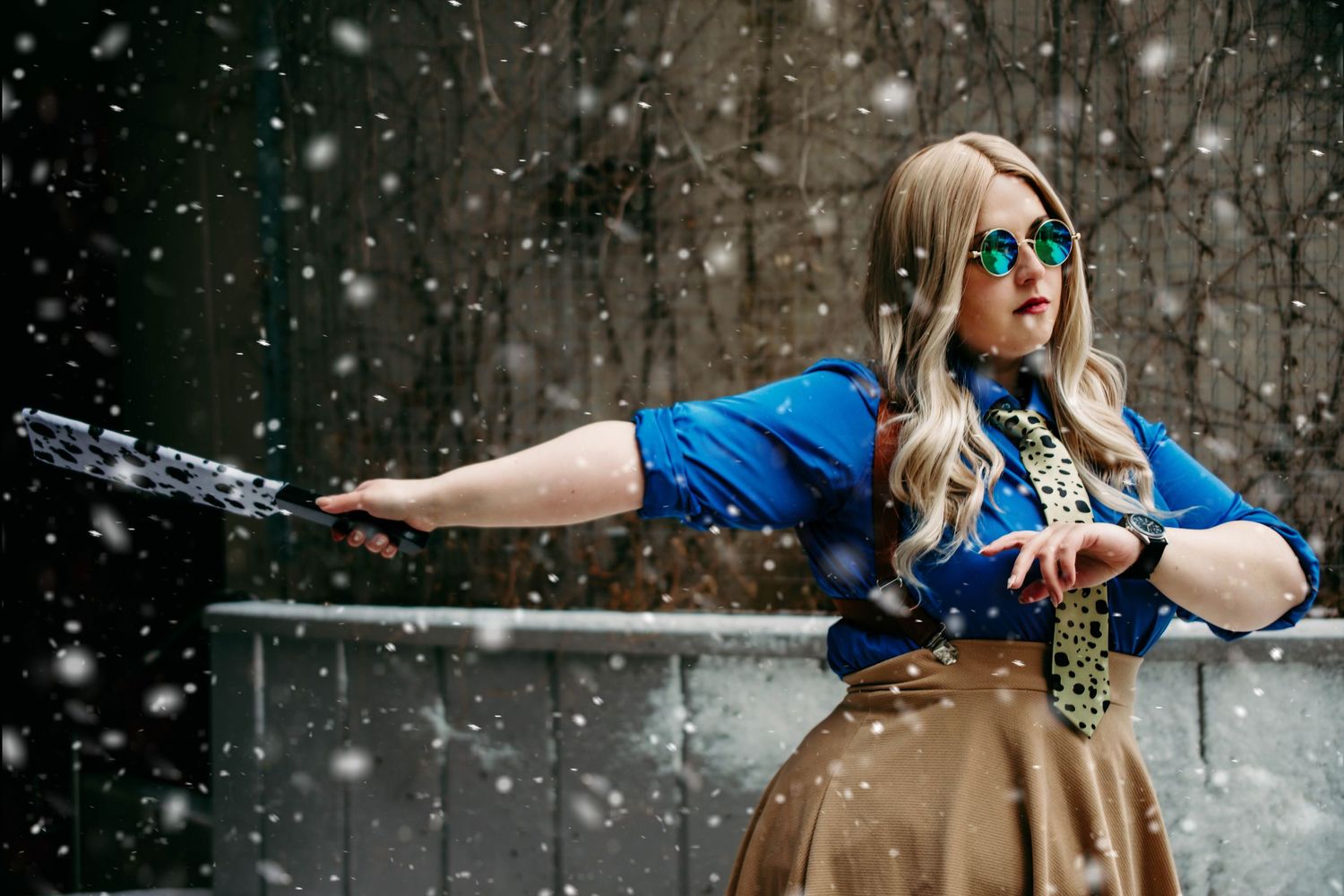 Winter cosplay photoshoot with falling snow and bright sunglasses against stone wall.
