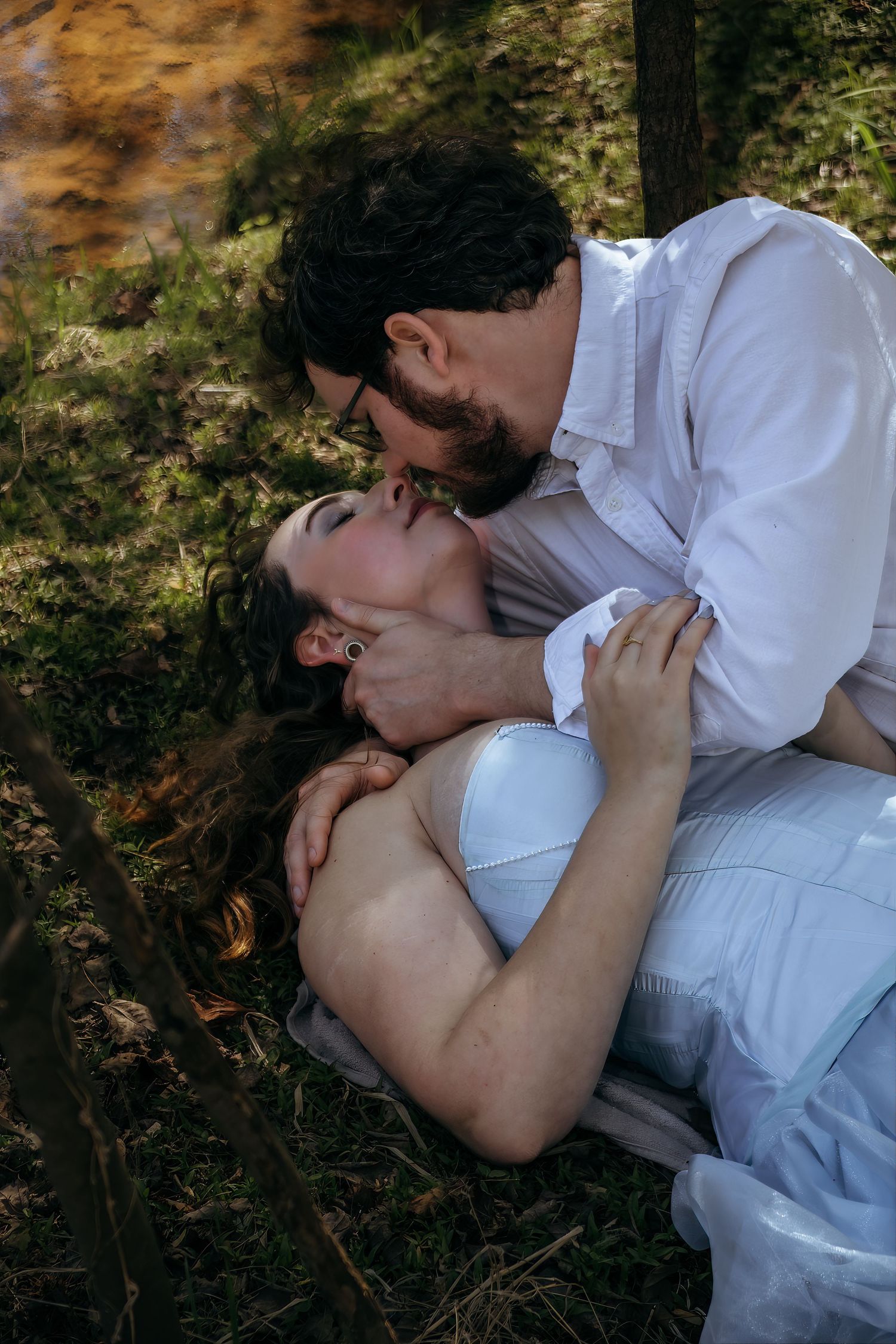 A couple shares a romantic moment outdoors, with the woman lying on grass and the man leaning in for a kiss.