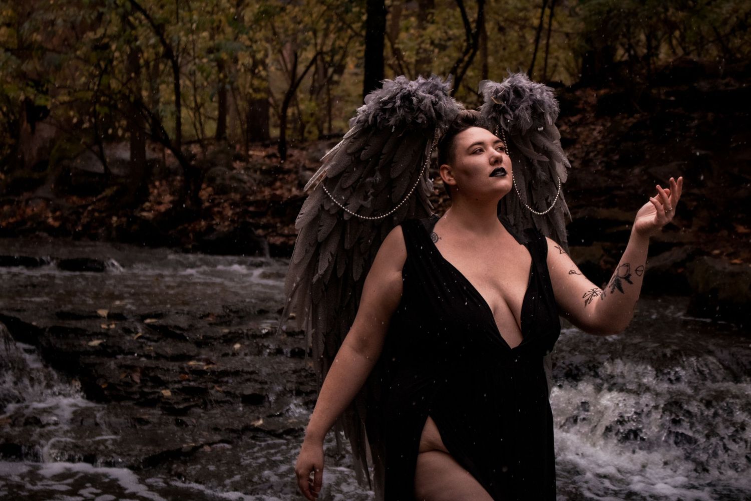 A figure in a black dress poses dramatically against a cascading waterfall in a moody forest setting.