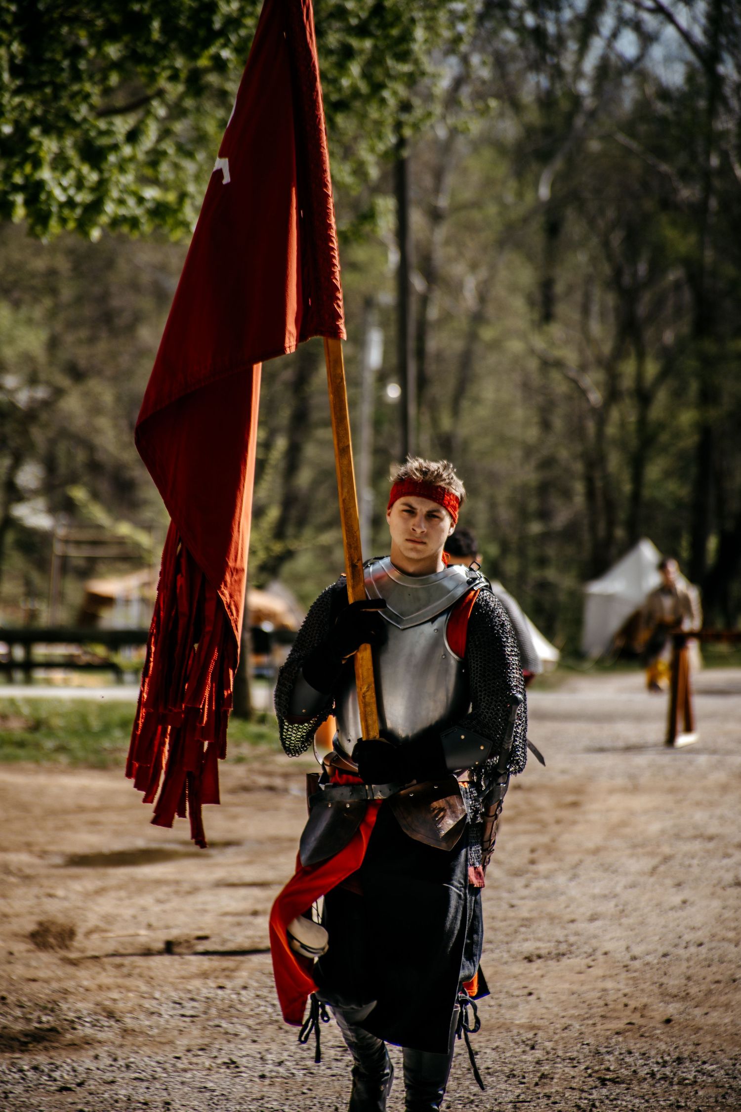 Young knight in medieval armor carries a red banner flag at an outdoor historical reenactment event in a forest setting.
