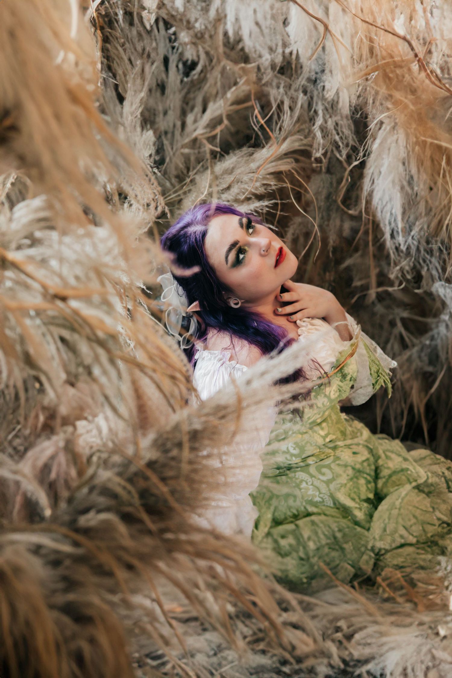 Woman with purple hair in green gown lying among dried pampas grass in artistic outdoor portrait.