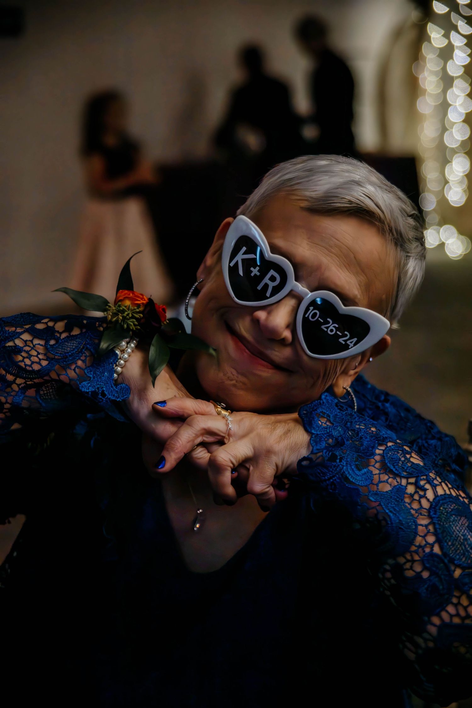 Woman in blue lace dress wearing heart-shaped wedding sunglasses with initials and date 10-26-24 at a wedding reception.