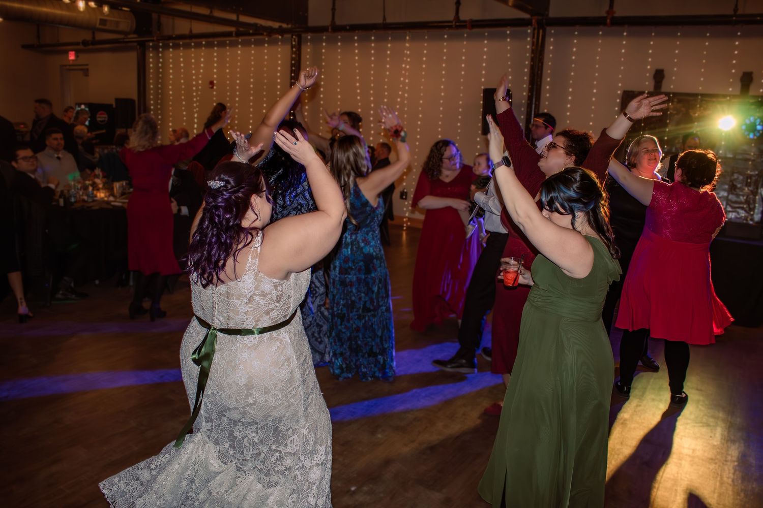 Bride and guests dancing joyfully at a wedding reception in a decorated venue with warm string lights.