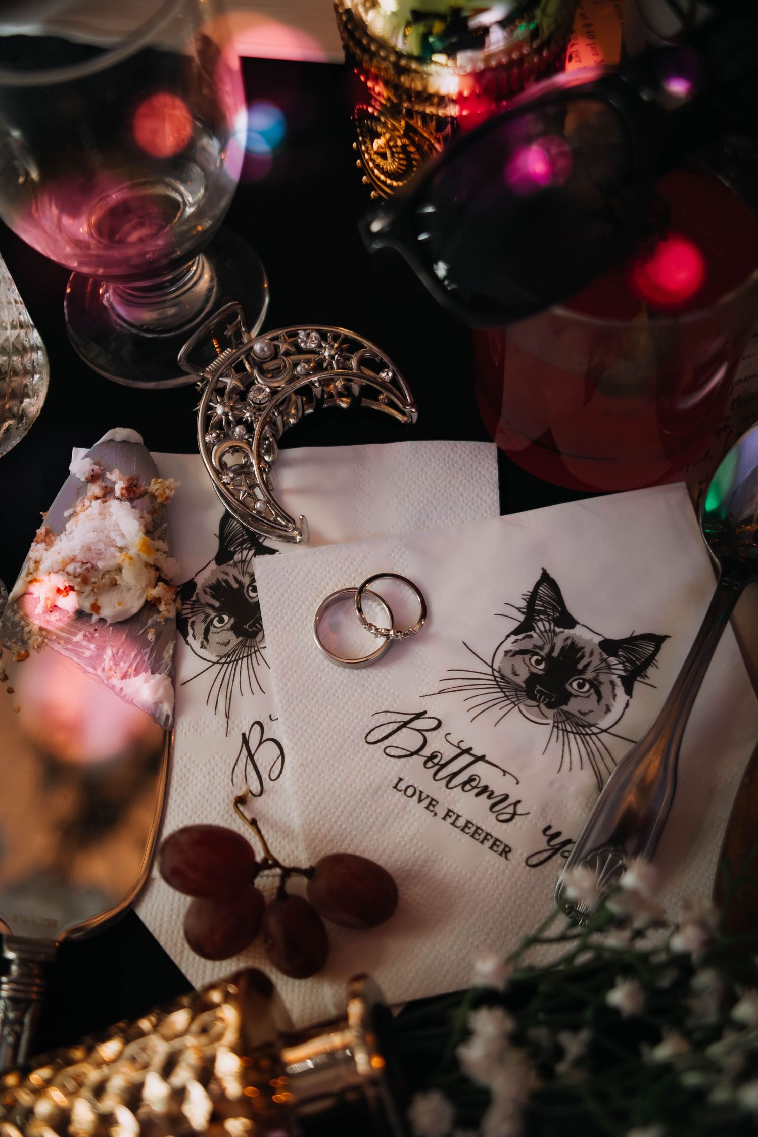 Wedding rings on custom cat napkin reading Buttons ye, with crescent moon brooch, grapes, and colorful bokeh lights.