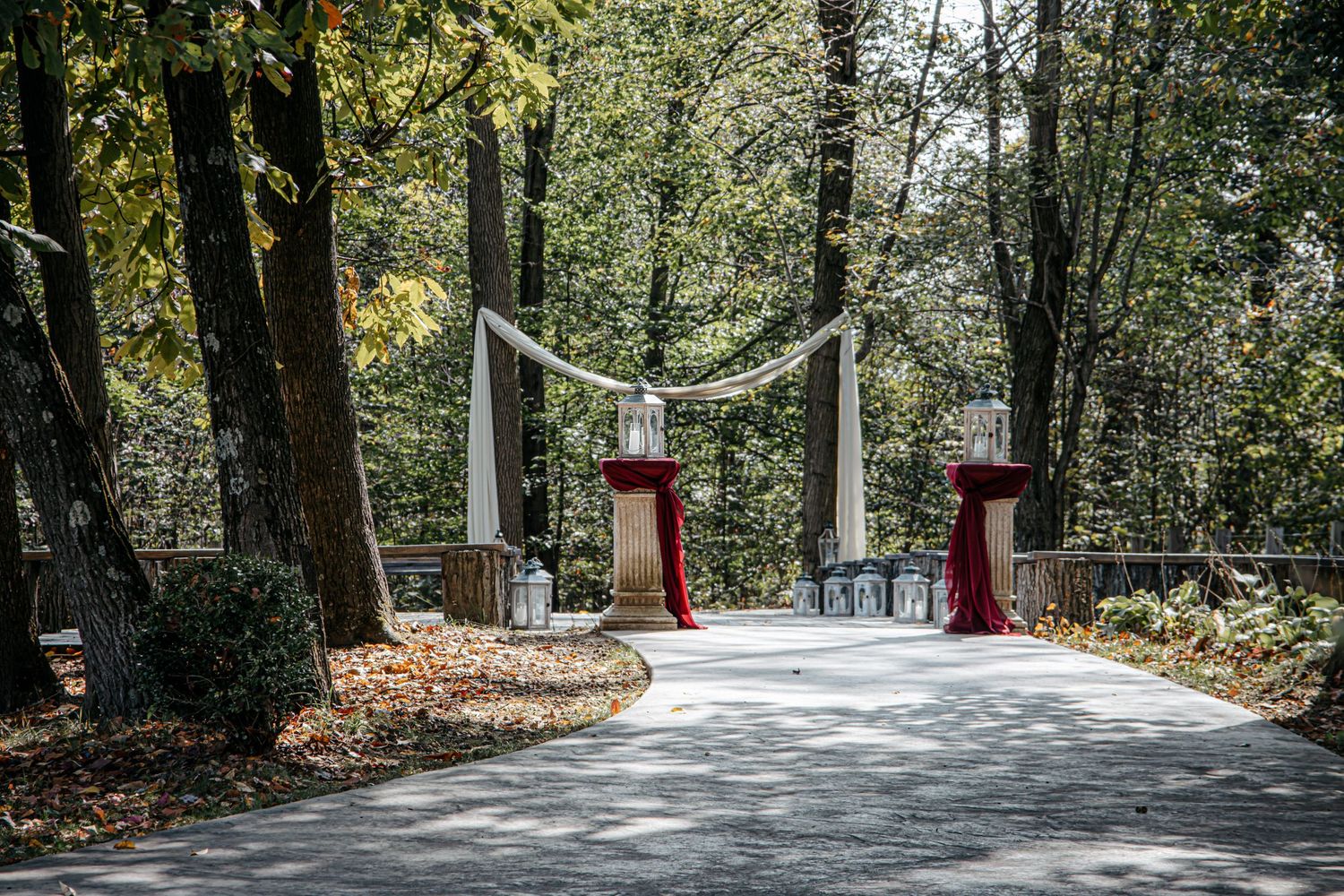 Elegant outdoor wedding entrance with red pedestal stands, draped white fabric, and lanterns on a tree-lined path.