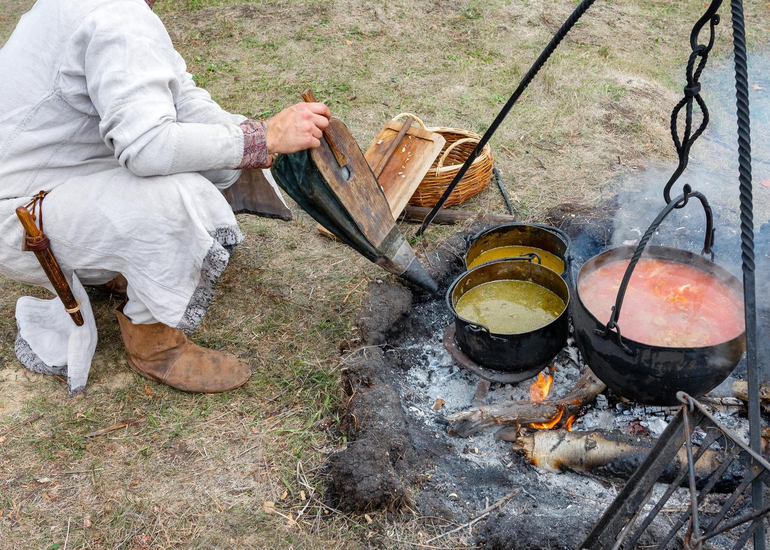Two cast iron pots hang over a campfire while someone tends to a wooden box nearby in an outdoor cooking scene.
