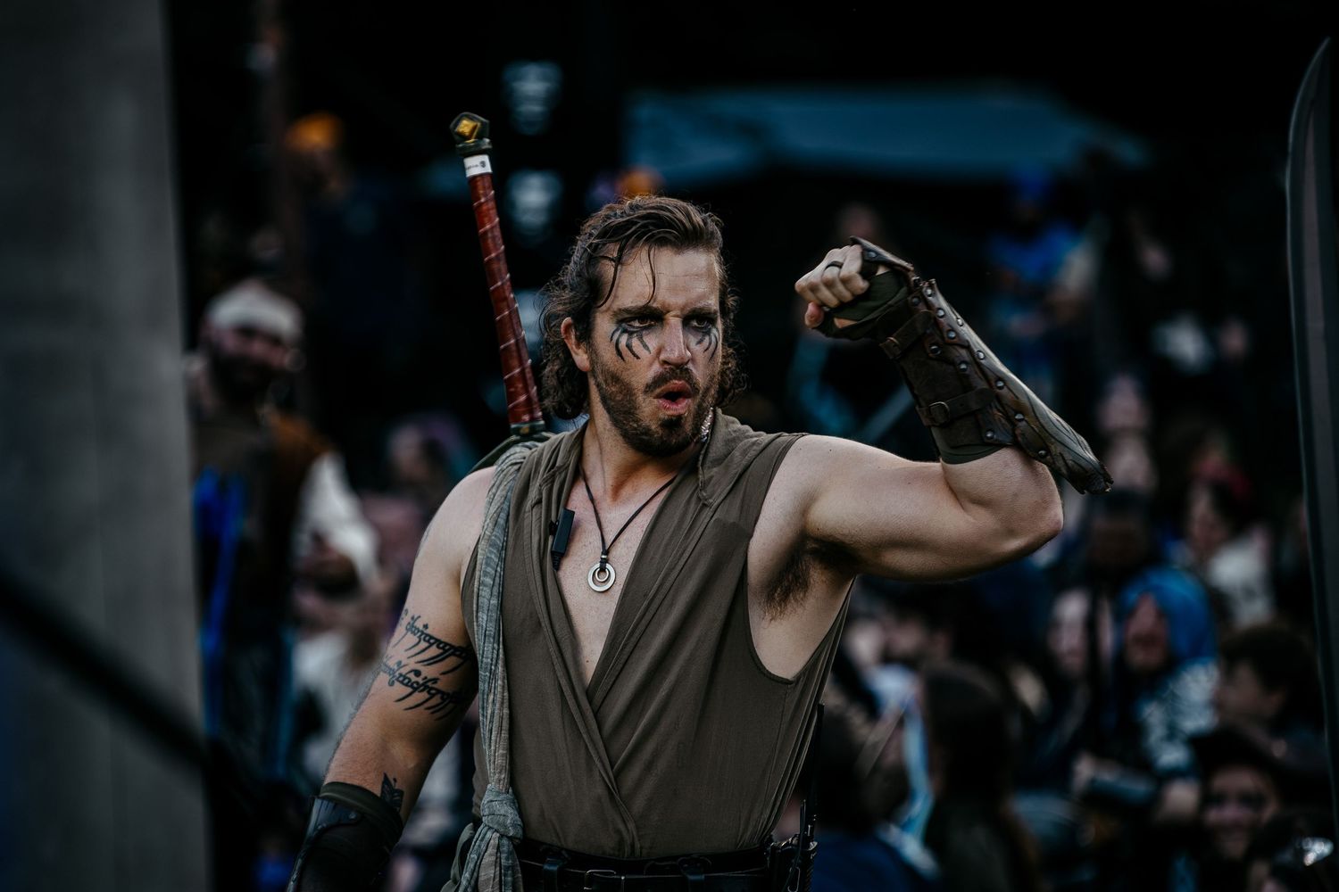 Warrior cosplayer with face paint, sword on back, raises fist dramatically at a crowded event.