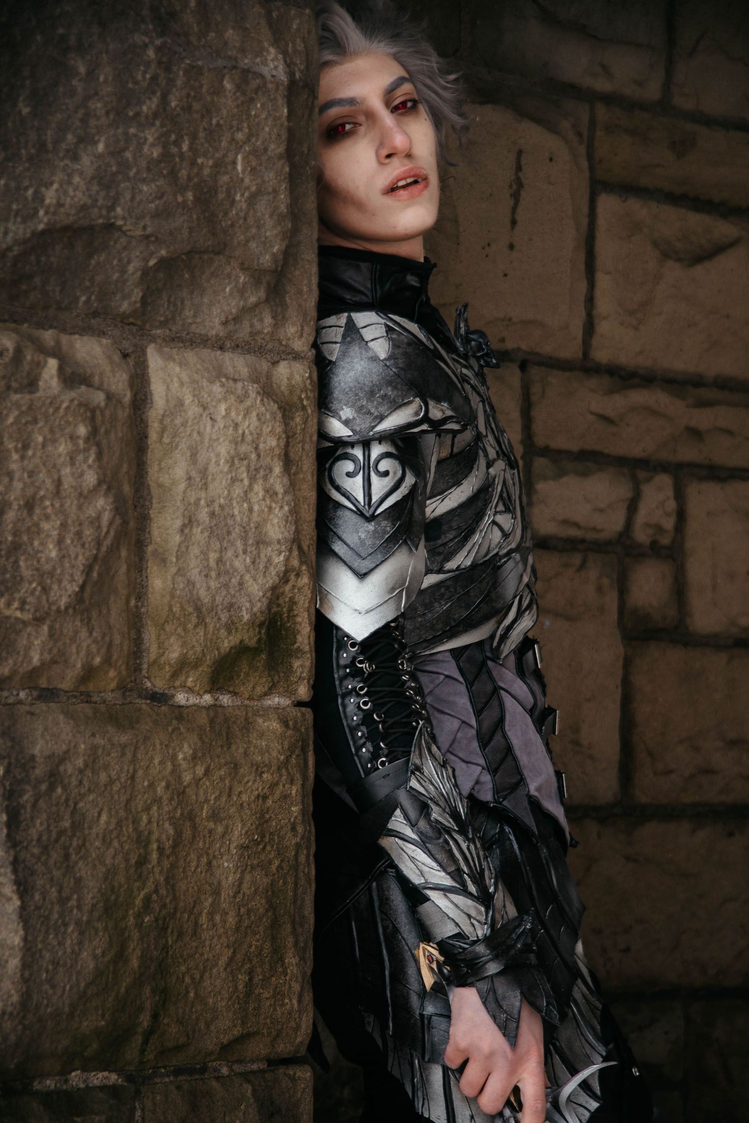 Cosplayer wearing metallic armor and dark clothing poses dramatically against stone wall in medieval setting.