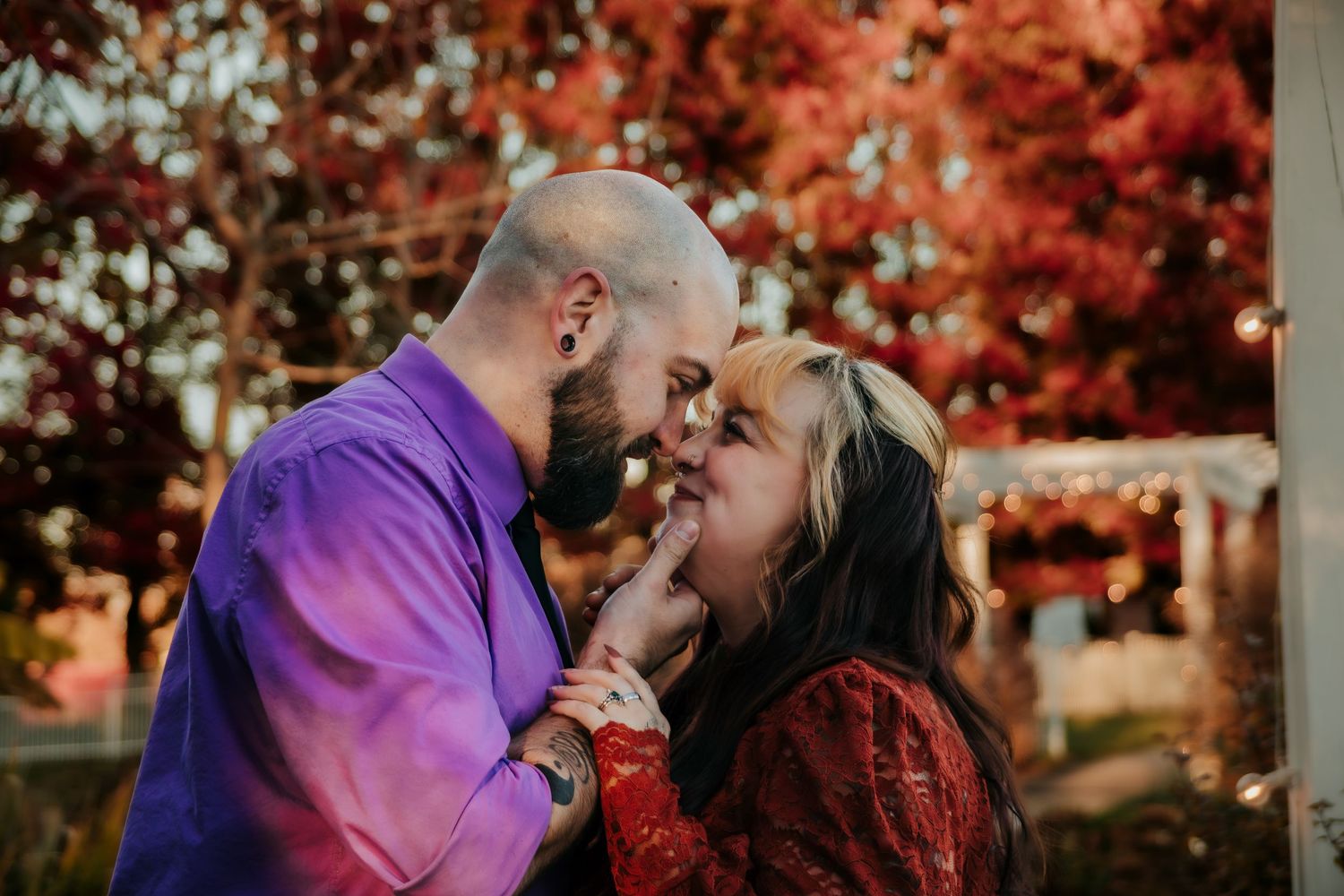 A couple shares romantic moments during an autumn photoshoot with vibrant red foliage in the background.