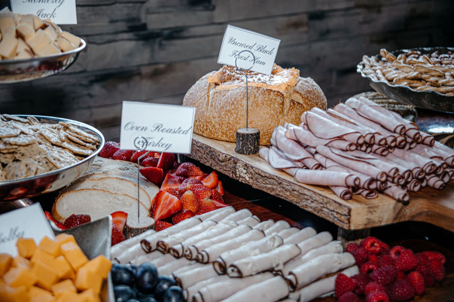 Assorted party platters with fresh fruits, meats, cheeses, and breads arranged on a rustic wooden table.