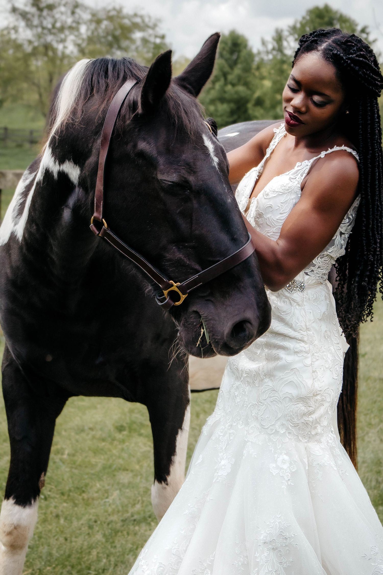 A bride in a white lace gown shares a tender moment with a black and white horse in an outdoor setting.