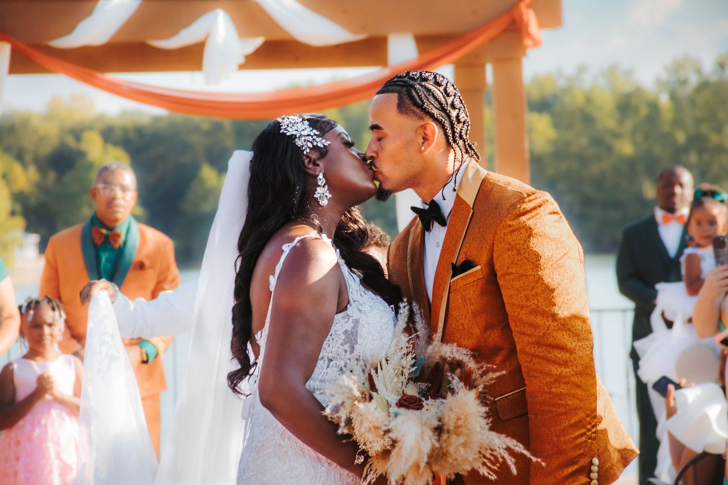 A couple shares a romantic kiss under an orange draped canopy during their outdoor wedding ceremony.