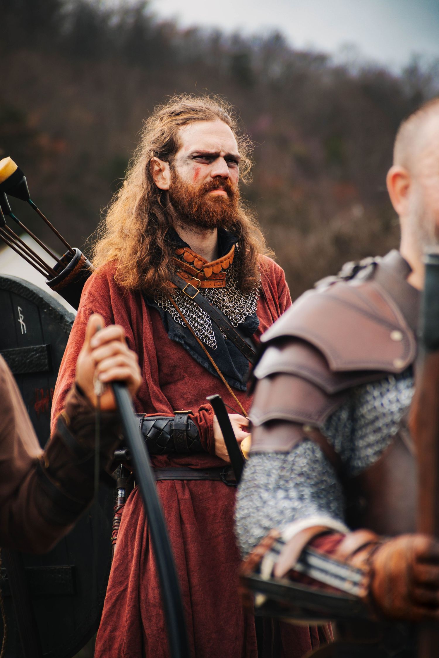 Medieval reenactor in red cloak with long red beard interacts with armored warrior during historical event.