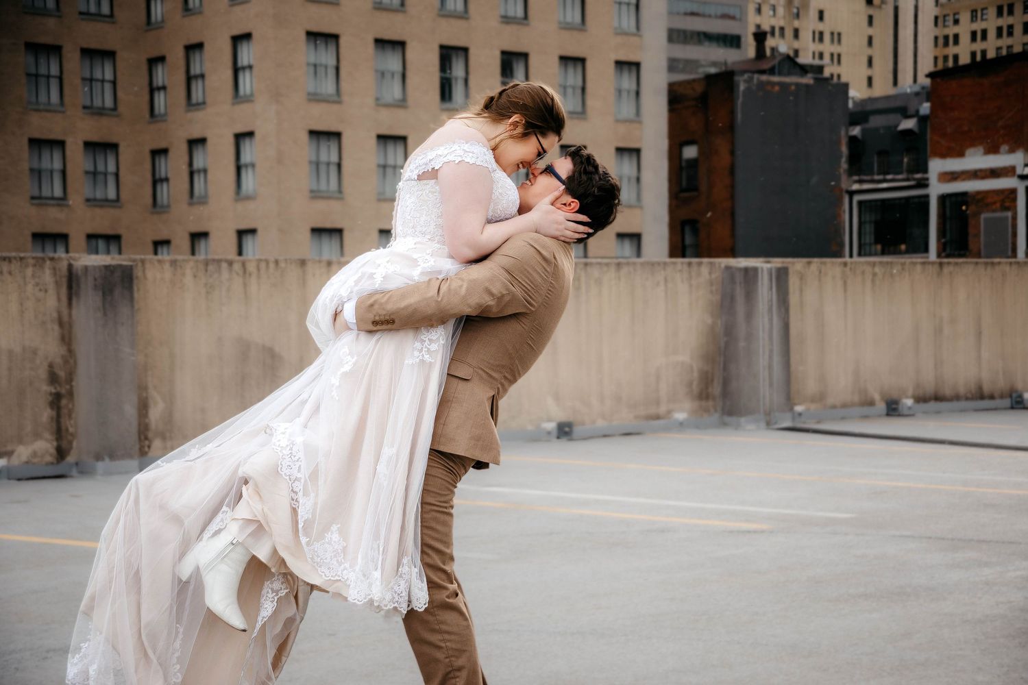 A couple in wedding attire embraces on an urban rooftop with city buildings in the background.