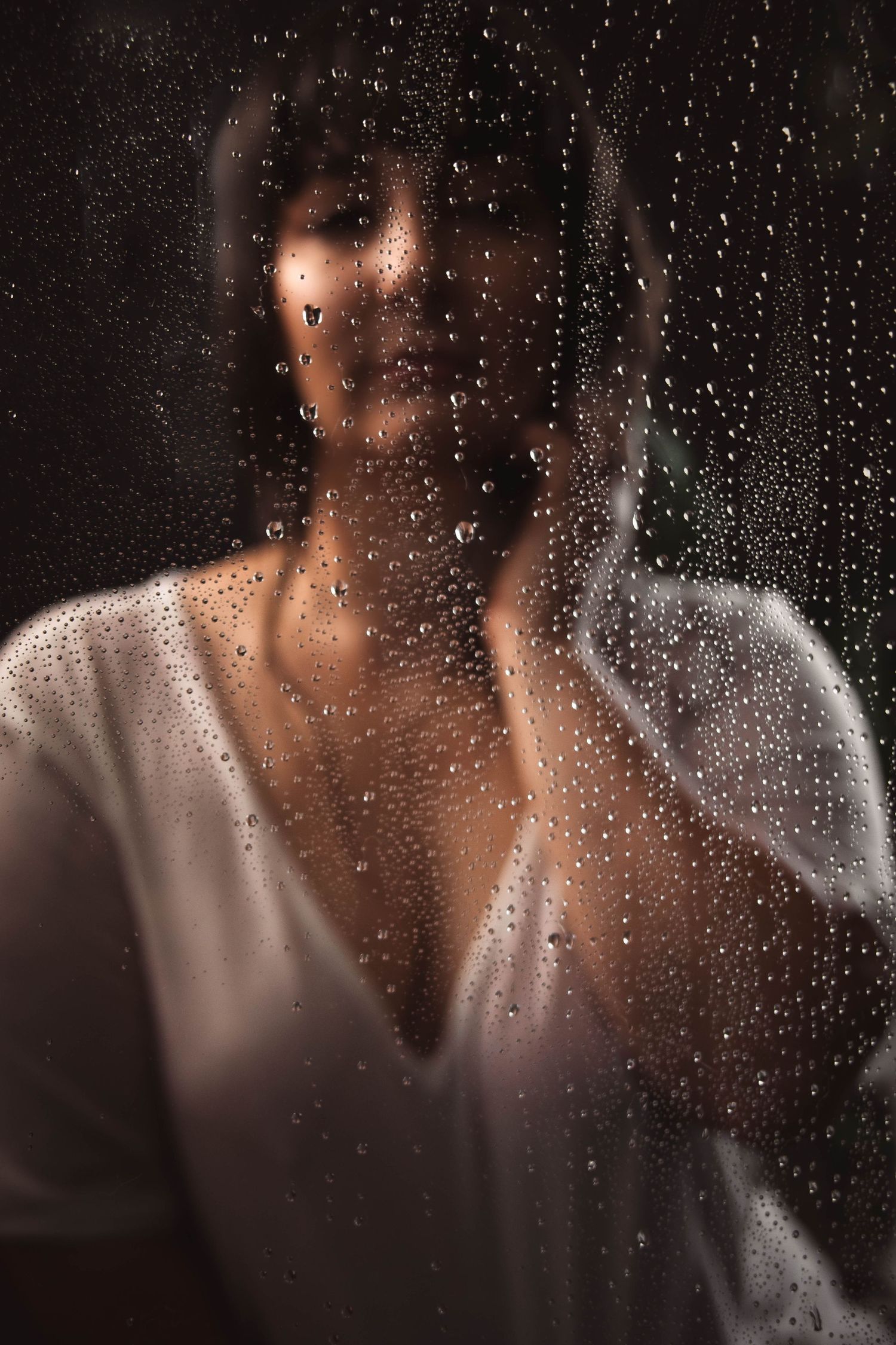 Artistic portrait through rain-covered window with soft focus and water droplets.