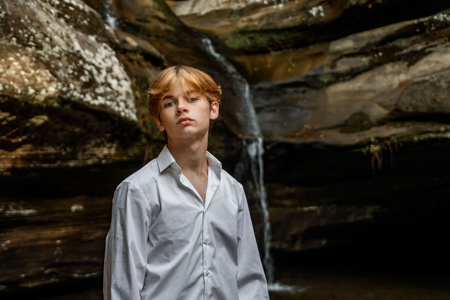 A person in a white shirt poses thoughtfully near rocky cave formations outdoors.