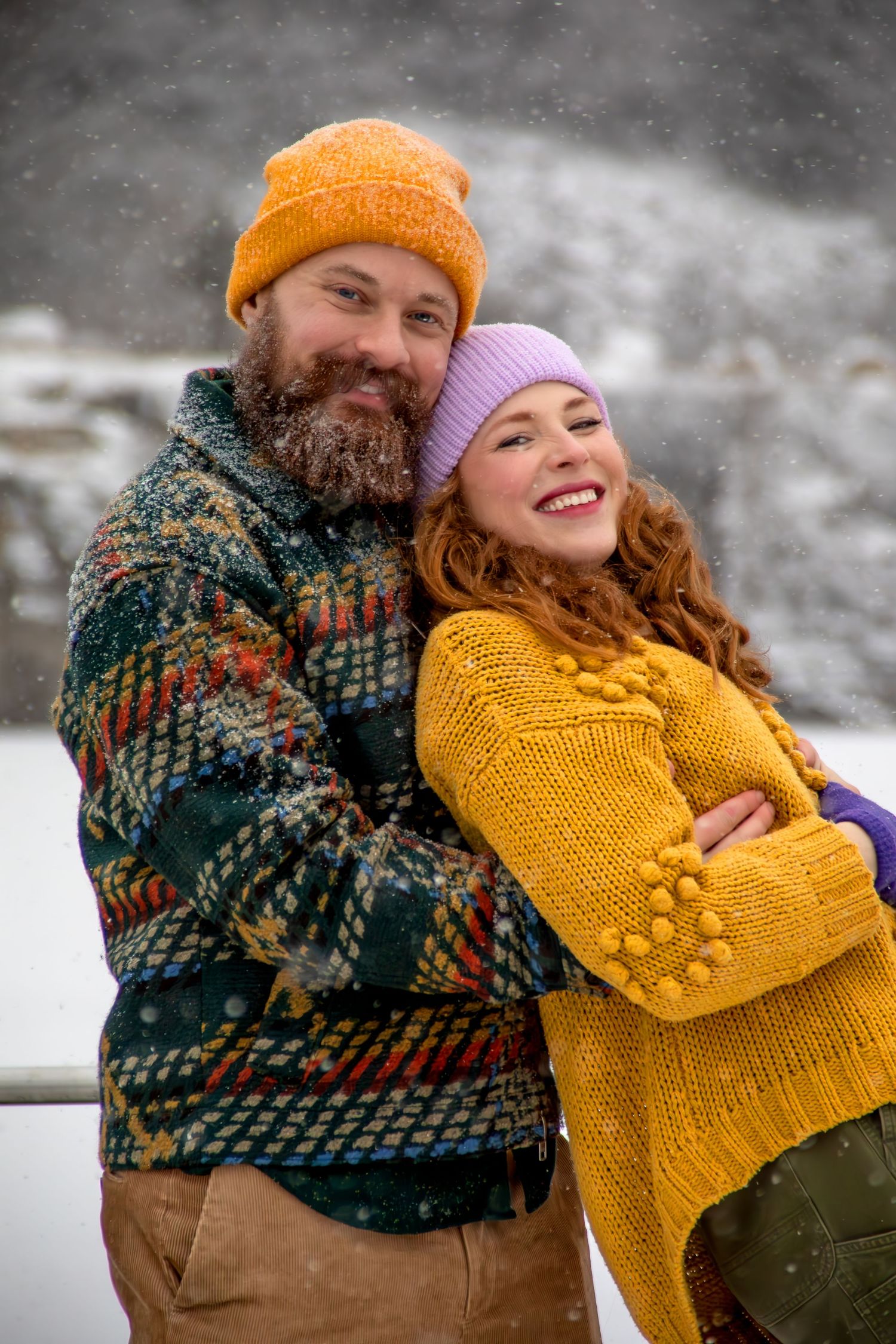A couple wearing winter clothing embraces and laughs together in the snow while wearing knit sweaters and beanies.