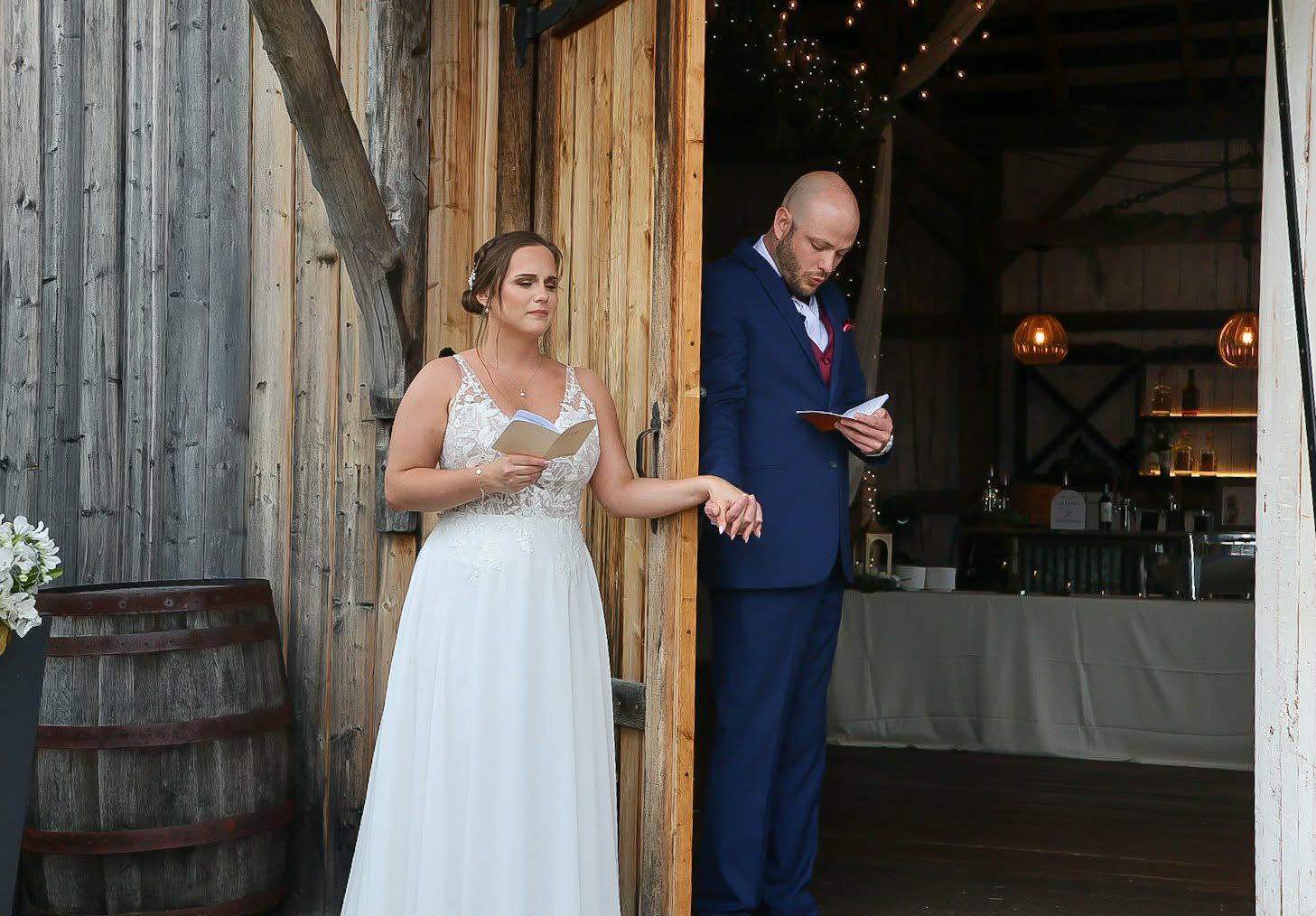 Bride and groom reading wedding vows at rustic barn venue, bride in lace gown, groom in navy suit.
