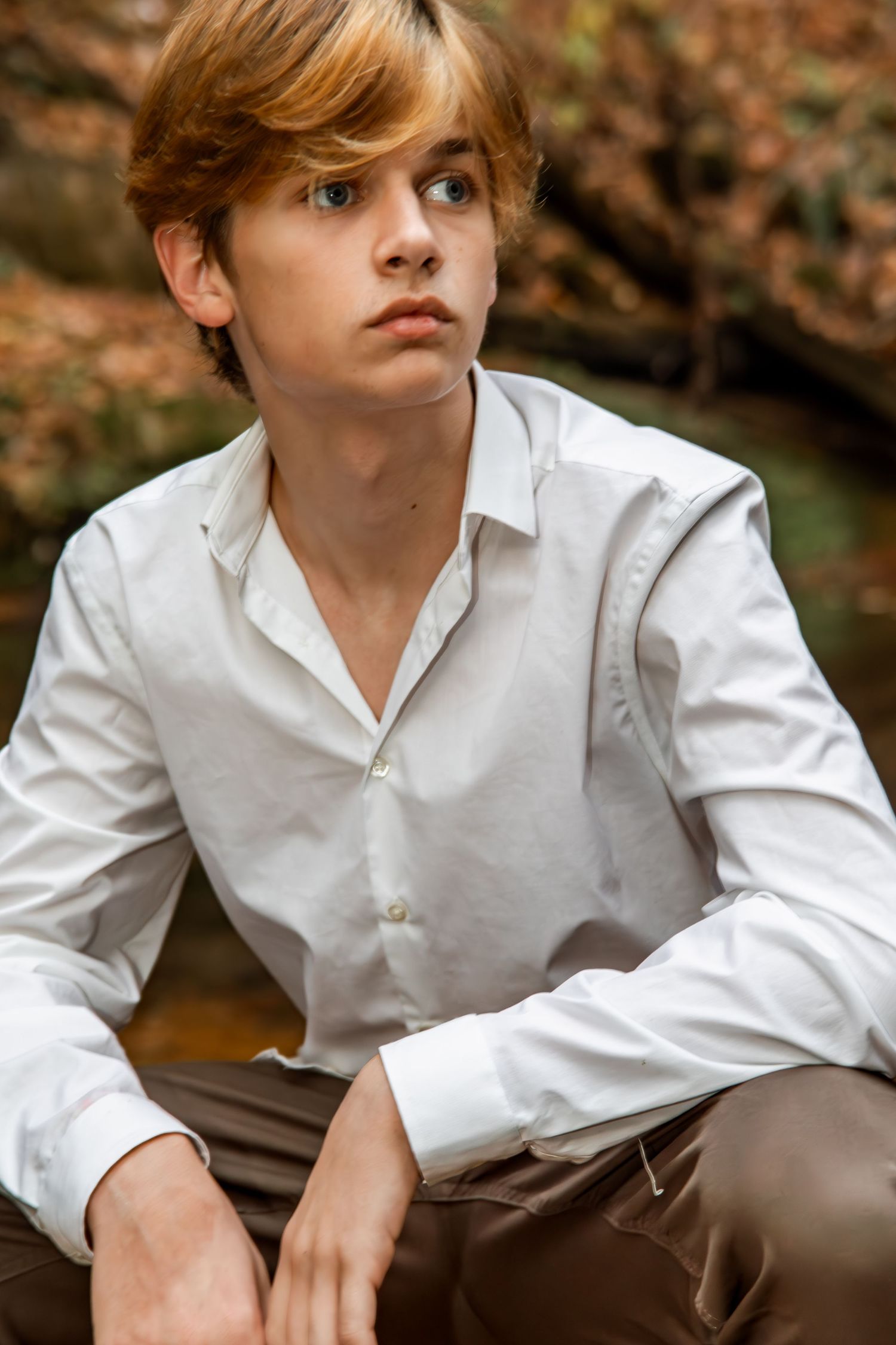 A young man with auburn hair in a white button-up shirt sits outdoors among autumn foliage, gazing thoughtfully to the side.