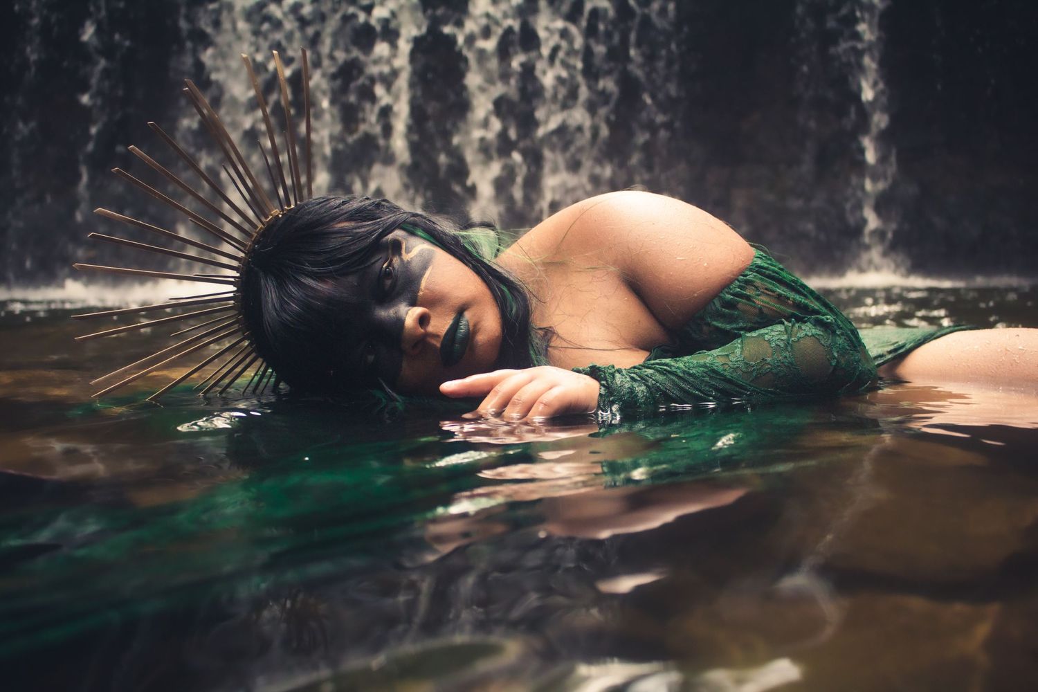 A woman with feathered headdress lies in shallow water before a waterfall, wearing a green lace outfit.