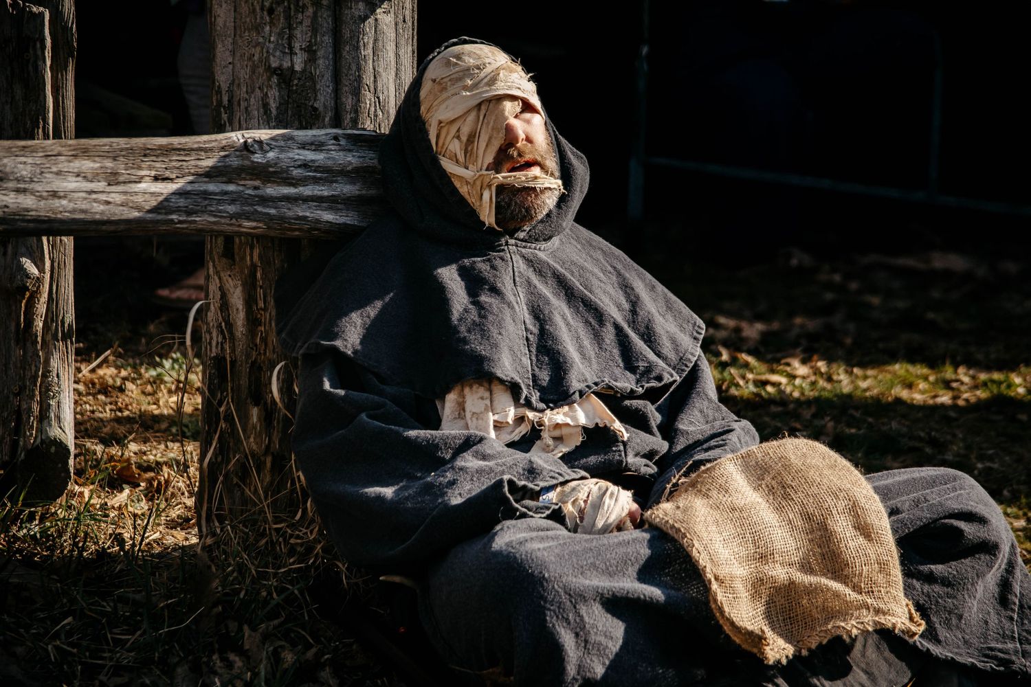 A weathered scarecrow figure in dark robes sits against a wooden bench outdoors in autumn light.