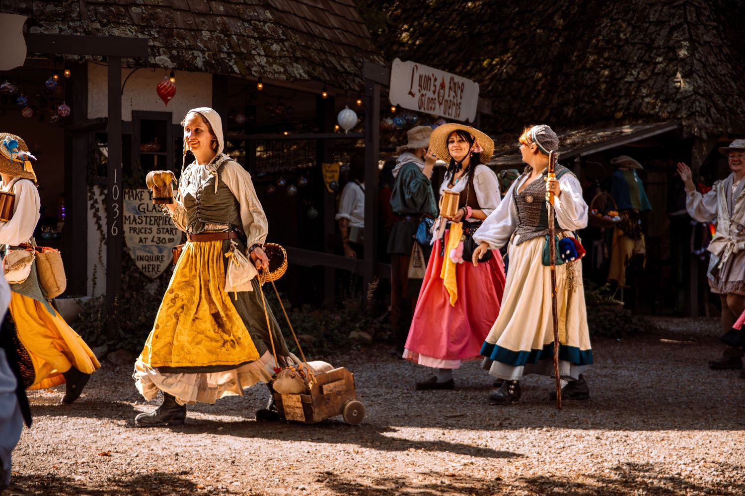 Women in colorful traditional folk costumes walk through a medieval market fair in golden sunlight.