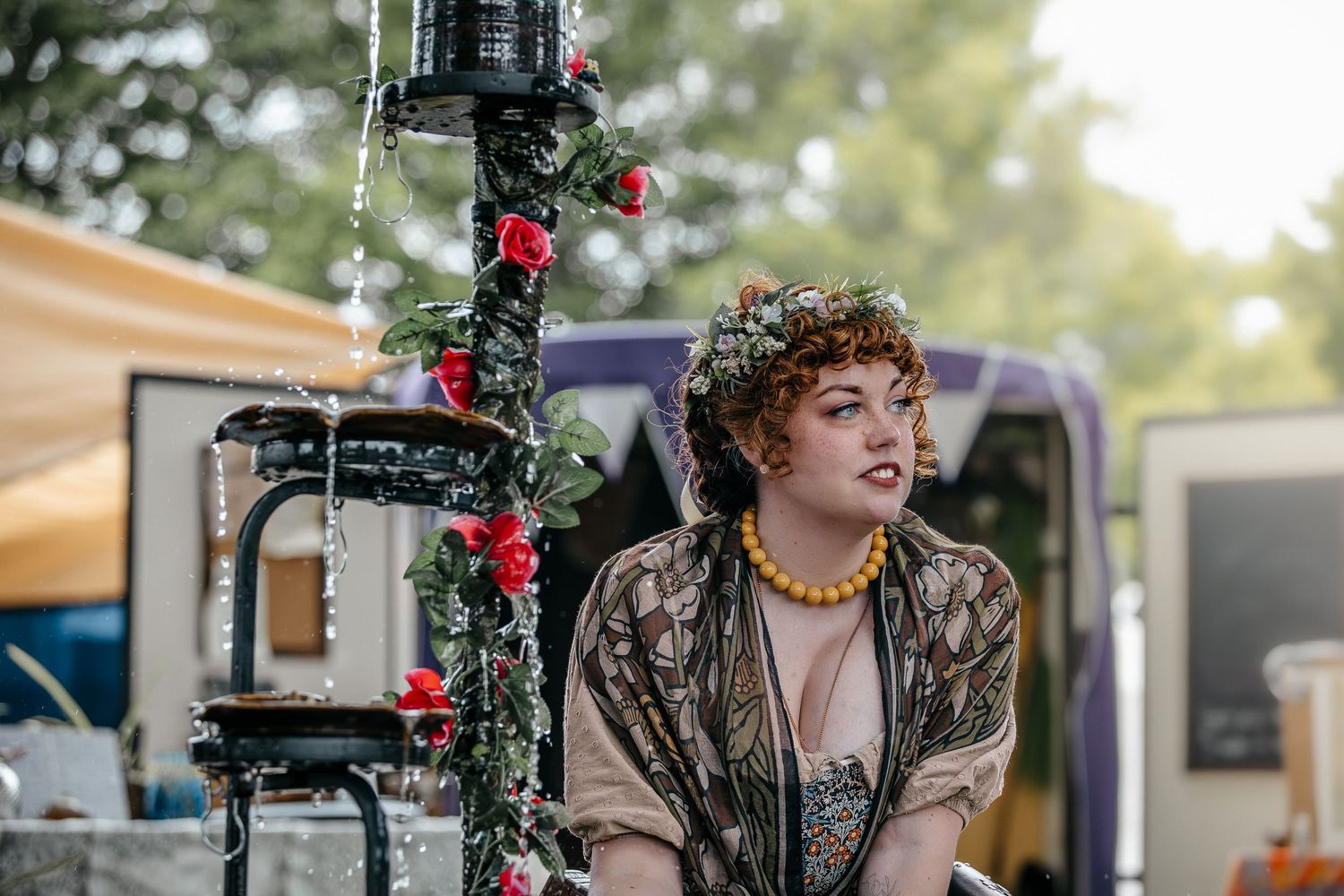 Woman with floral crown and yellow necklace poses near a decorated pole with red roses at an outdoor festival.