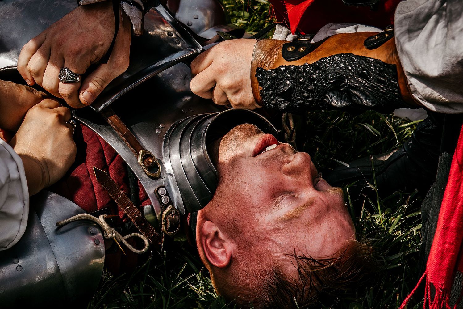 Medieval reenactment scene showing a fallen warrior in armor lying on grass with a sword held to his throat.