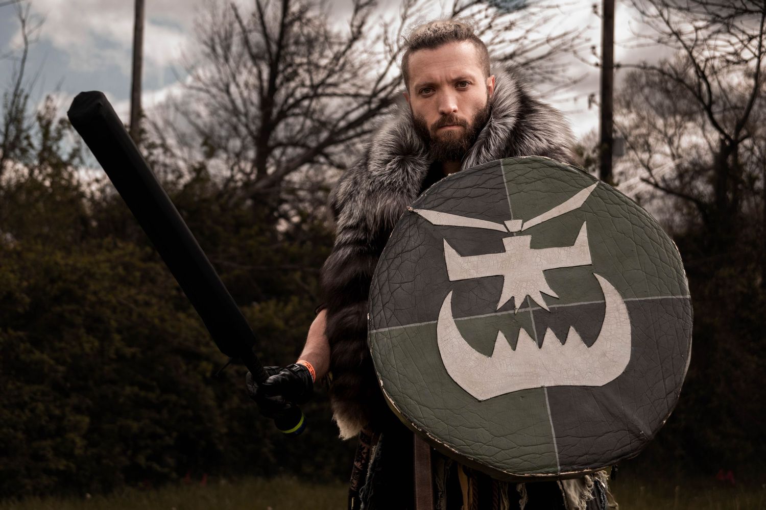 A person in medieval costume holds a round shield with a stylized design against a dark forest backdrop.
