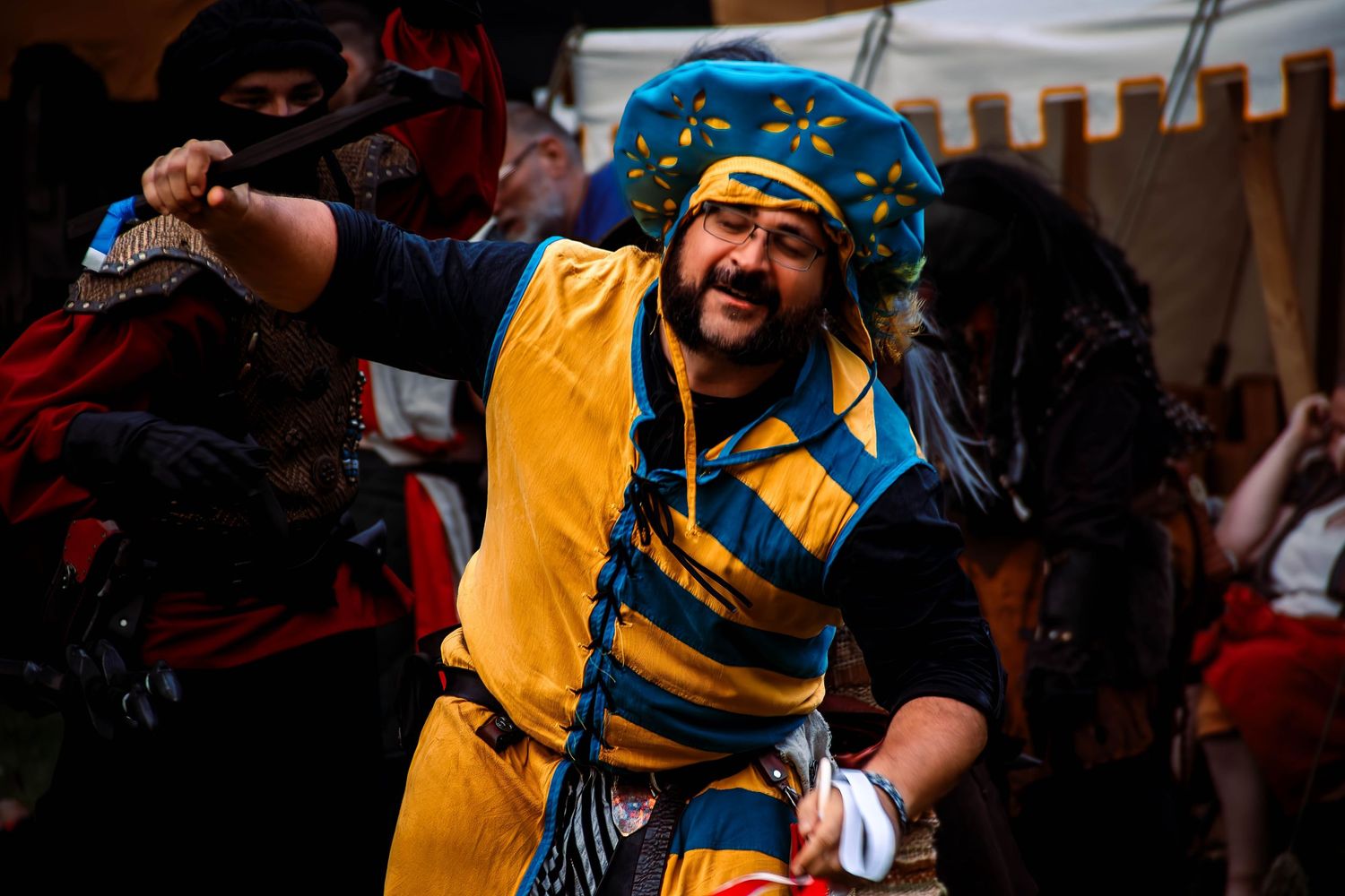 A performer in a bright yellow and blue striped medieval costume smiles enthusiastically during a festival performance.