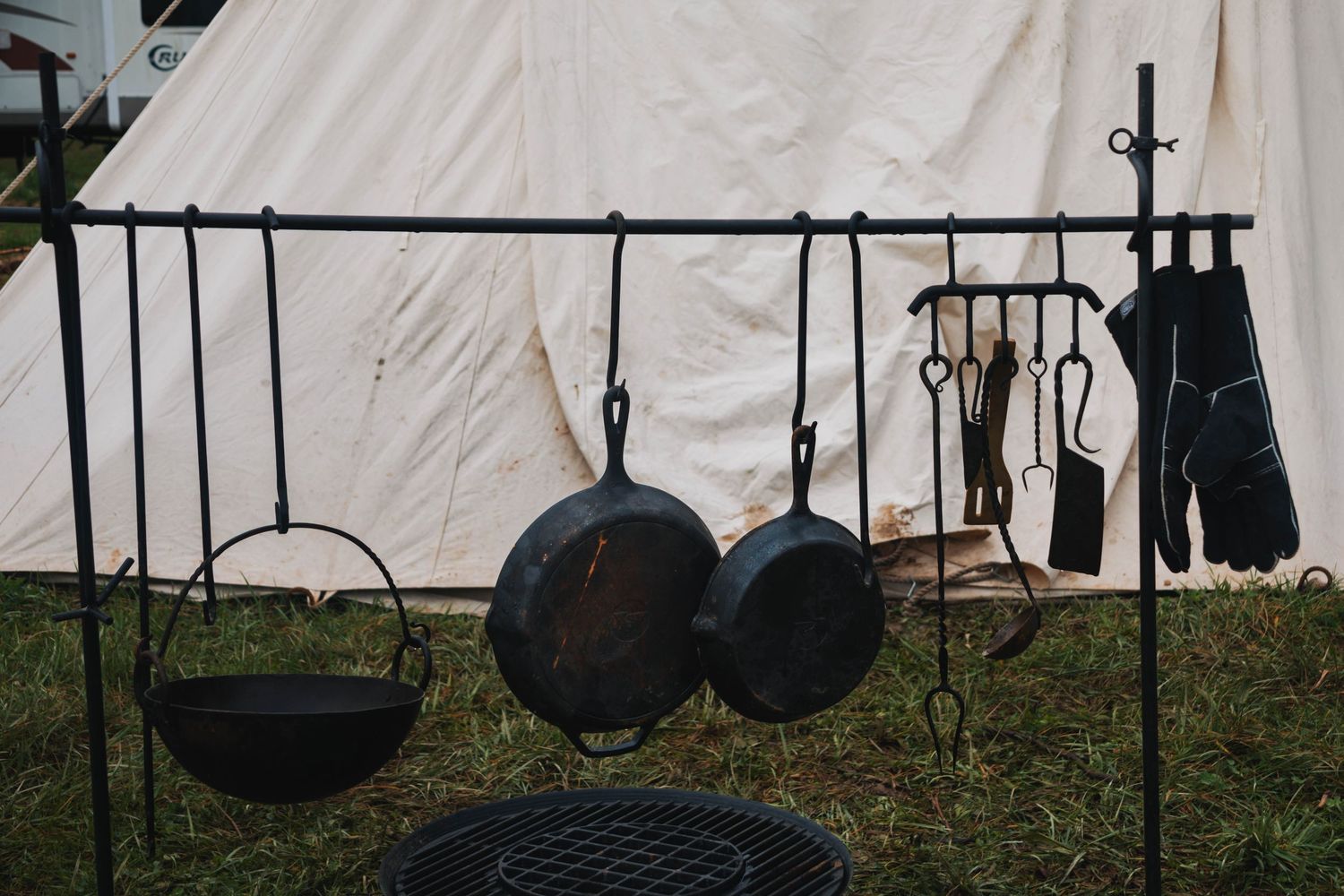 Rustic black cast iron pots and pans hanging from hooks against a white canvas tent background at an outdoor event.