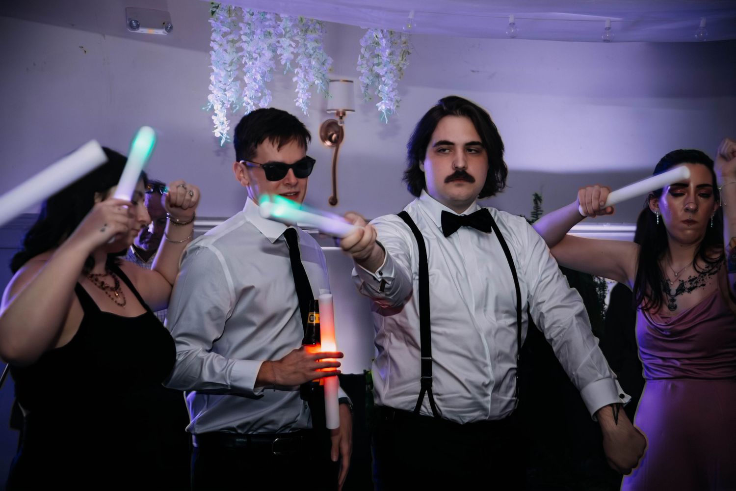 People dancing at a wedding reception holding glowing foam sticks, men in suspenders and bow ties celebrating.