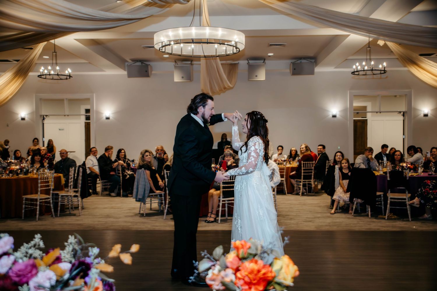 Bride and groom share first dance in elegant ballroom with chandelier, floral arrangements, and seated wedding guests.