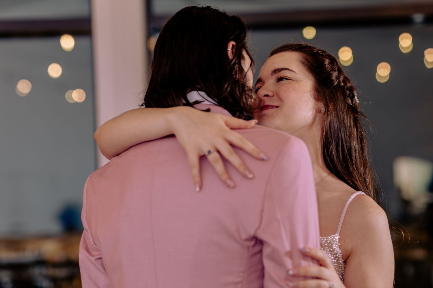 Couple sharing a romantic dance embrace, smiling joyfully at a wedding reception with warm bokeh lights.