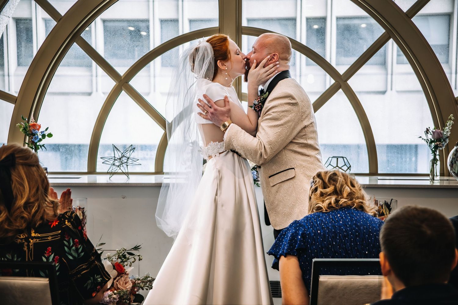 Bride and groom share first kiss at wedding ceremony before large arched window, guests seated in foreground.