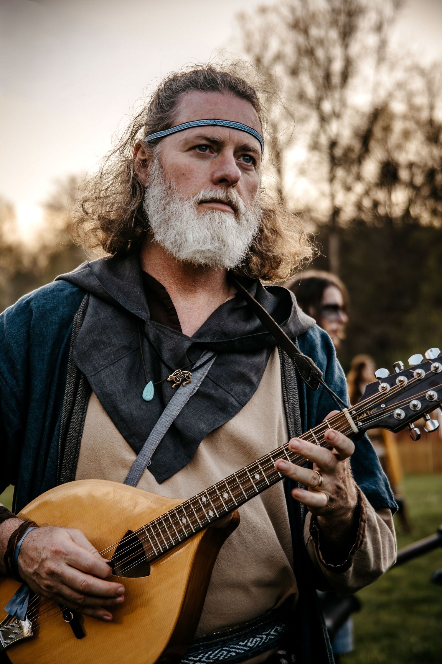 Older man with long hair and white beard playing a mandolin outdoors, wearing medieval-style clothing and a headband.