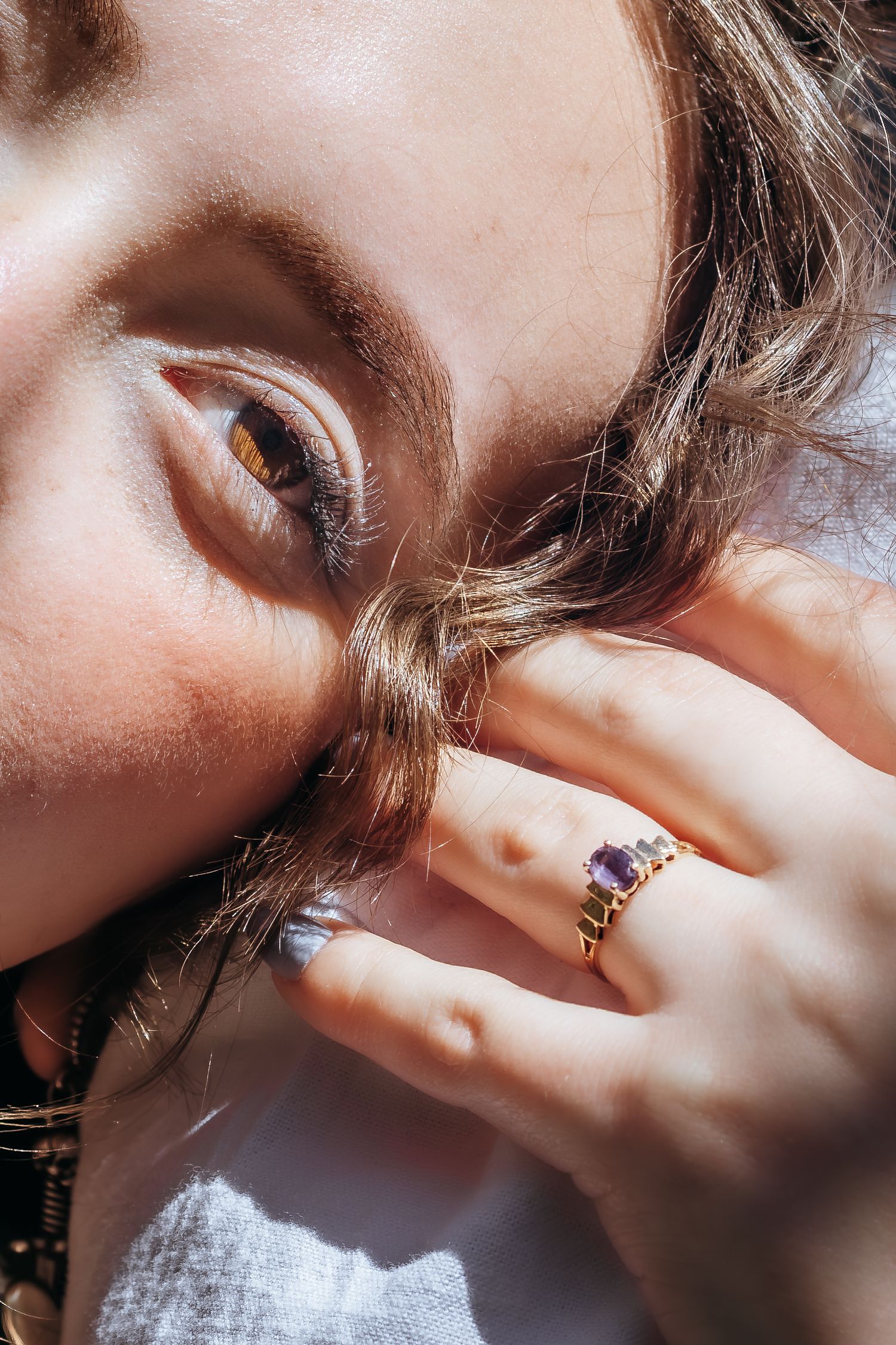 Close-up of a woman wearing a gold ring with purple gemstone, her hand resting near her face in soft sunlight.