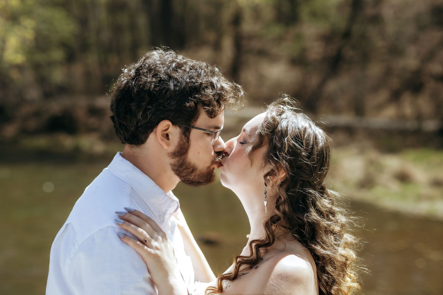 Couple sharing a romantic kiss outdoors near a river, surrounded by lush green trees in a natural forest setting.
