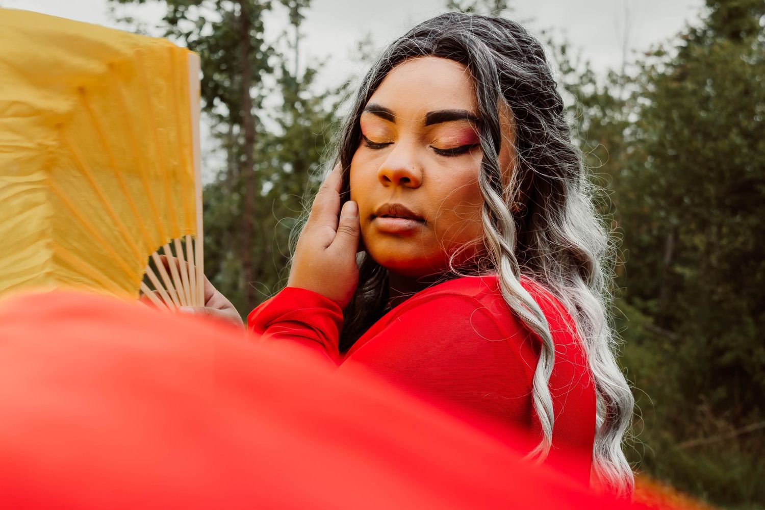 Woman in red outfit holding yellow fan with eyes closed, long gray braided hair, outdoors with green trees.
