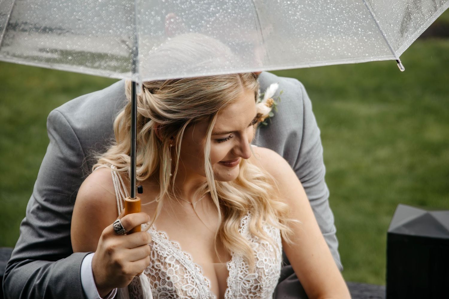Bride smiling under a clear umbrella in the rain, wearing a lace wedding dress with a groom in a gray suit beside her.
