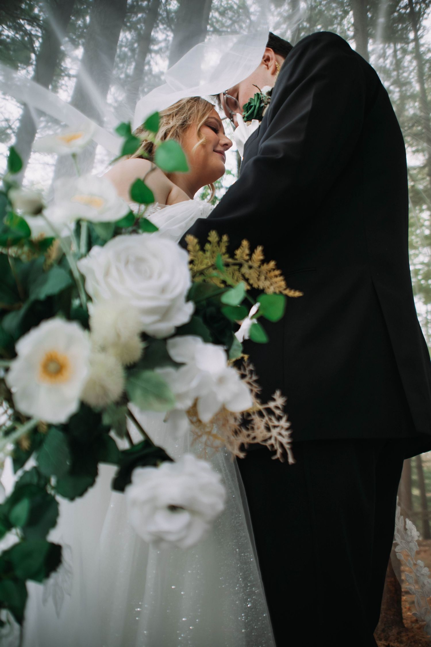 A romantic wedding couple shares tender moments in a misty forest setting with white floral bouquet details.