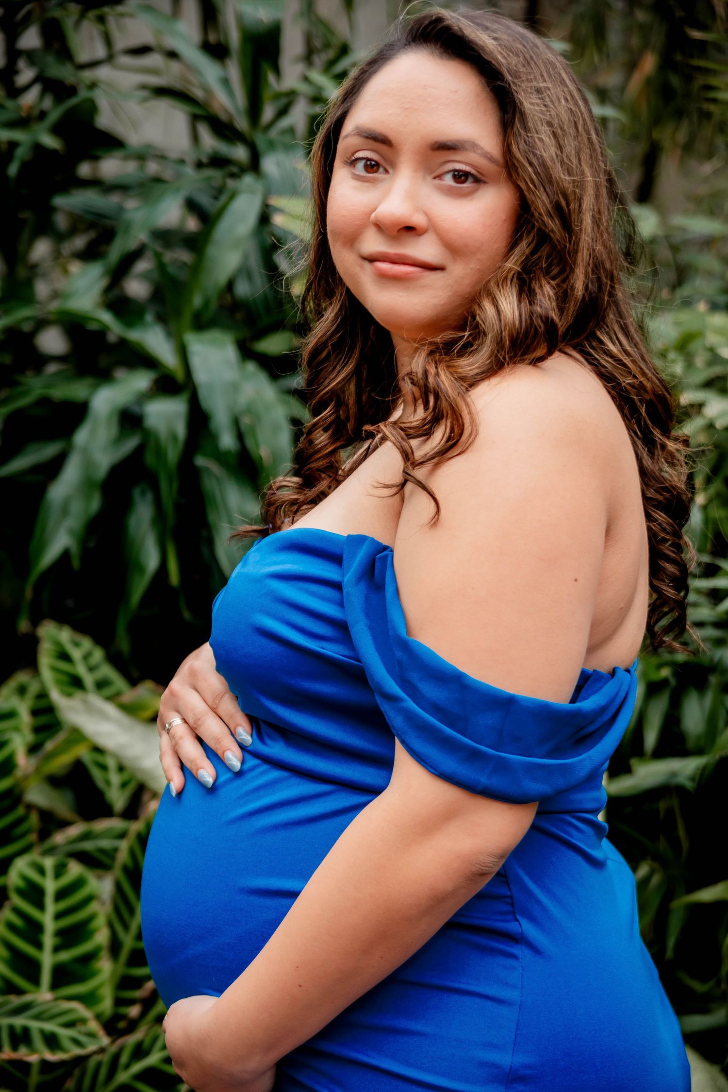 A maternity portrait featuring a woman in a royal blue off-shoulder dress against lush greenery.