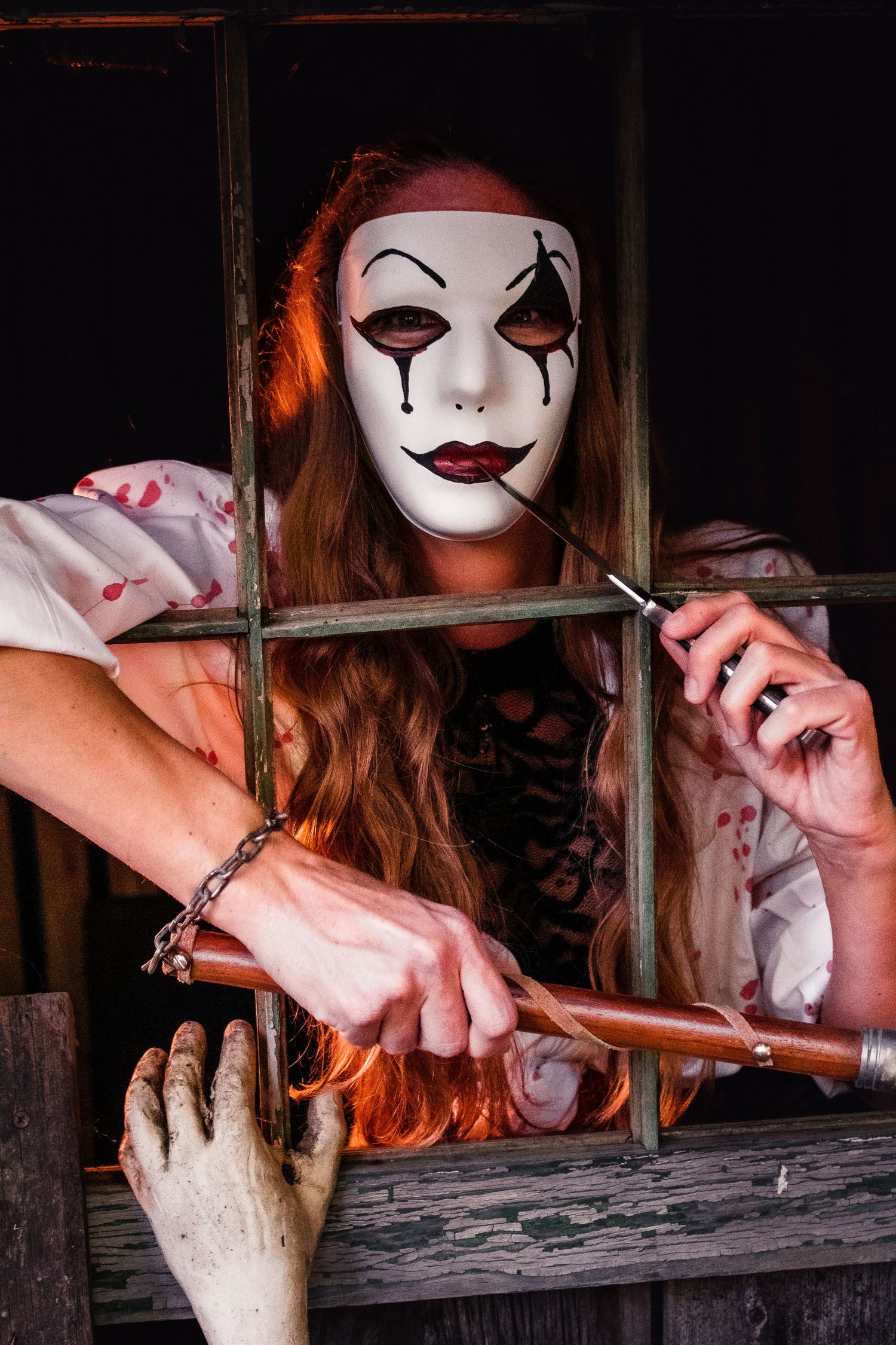 A creepy figure in white face paint and dark makeup reaches through wooden bars in a dark setting.