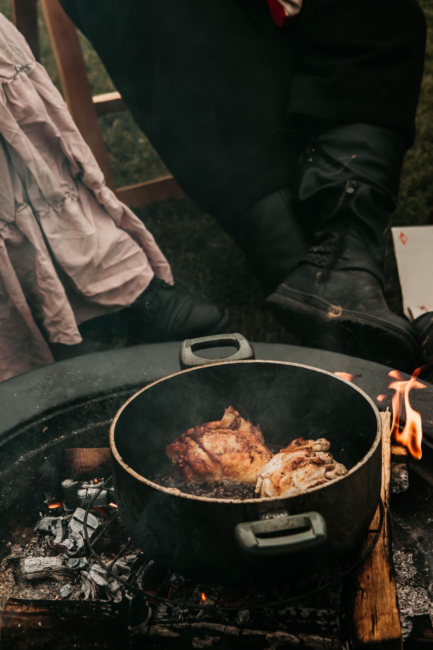 Meat cooking in a cast iron pan over open flames with boots visible in the background.