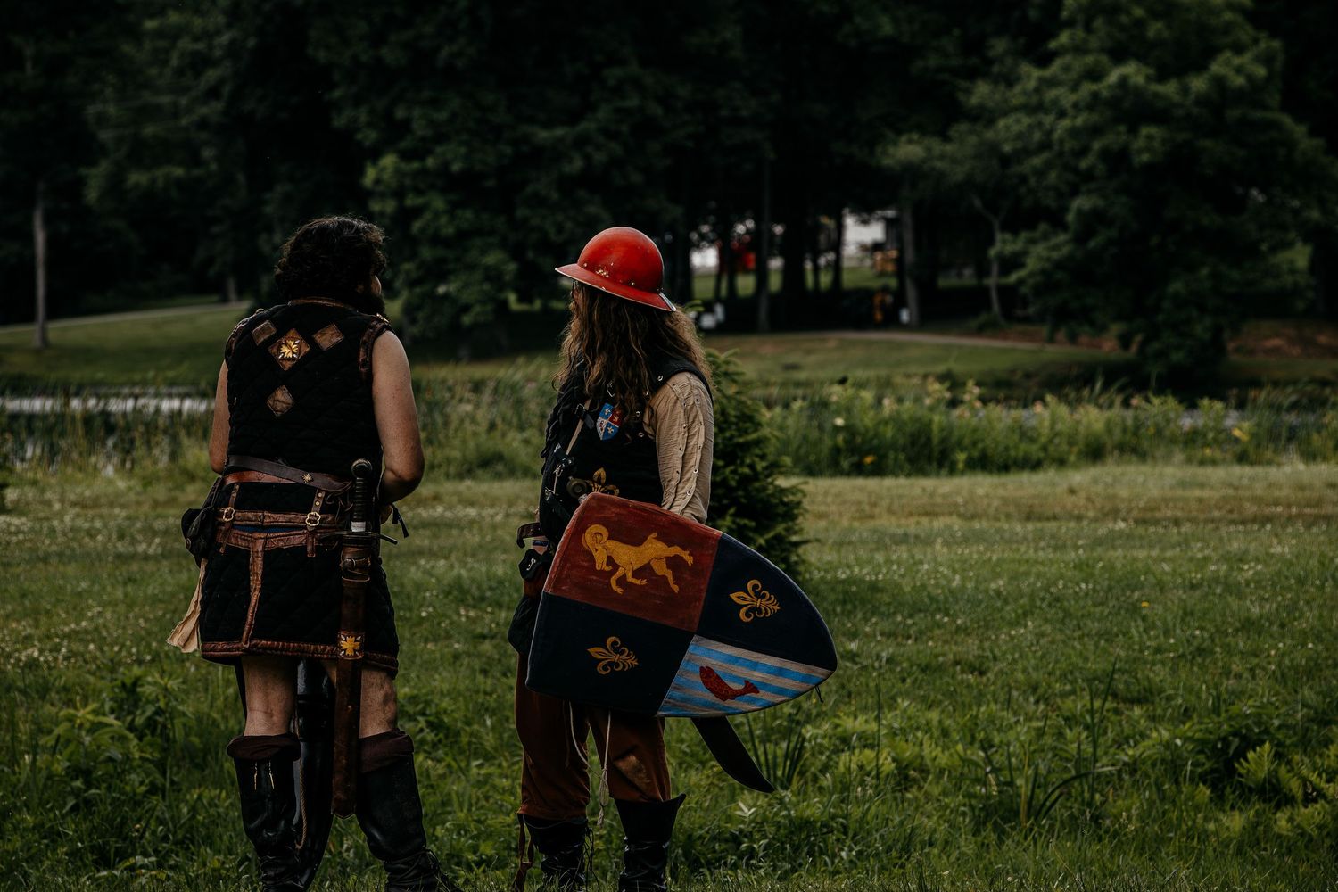Two people in medieval costumes carrying shields walk through green field at Renaissance festival.