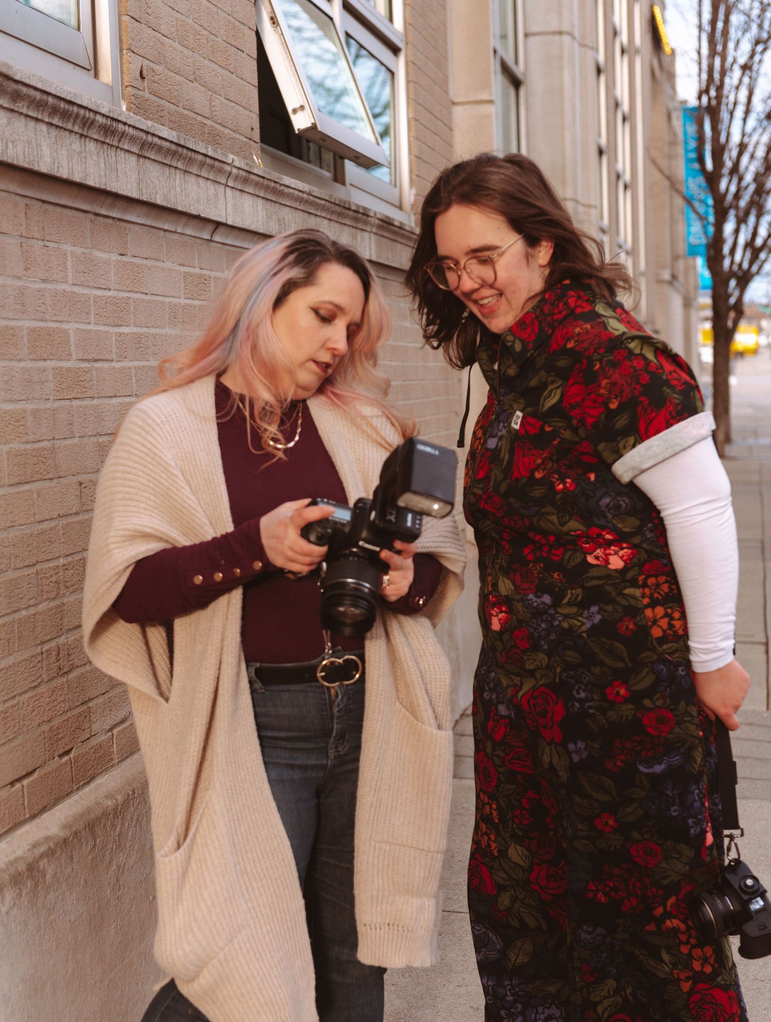 Two photographers in casual attire review images on a camera display against a brick wall background.