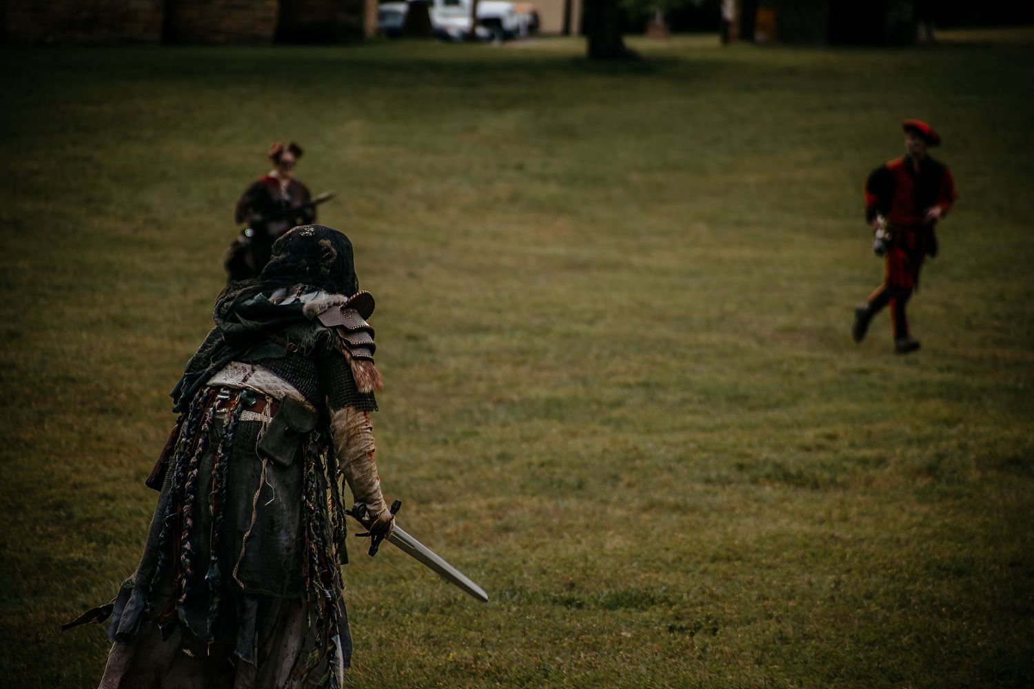 Medieval warriors in costume reenactment battle on green field, one holding sword facing opponent in red hat.